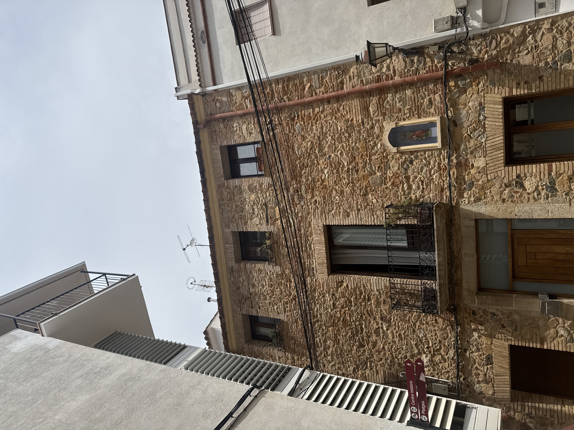 The image shows a view looking up at buildings in a narrow street in Tossa de Mar, Spain. The central building is made of rough-hewn stone with exposed brickwork around windows and doorways. It has a red-tiled roof and a small balcony with decorative iron railings. Above the balcony, there is a niche with a religious statue. Several windows are visible on the facade, some with small potted plants on their sills. Electrical wires are strung across the front of the building.
To the left of the stone building is a modern, white building with a textured facade and a protruding balcony with a metal railing. A set of horizontal louvers is visible on this building. A signpost with directional arrows indicating "Centre històric" and "Platges" is attached to the stone building.
To the right of the stone building is another building with a white facade and a scalloped decorative trim along the roofline. A black lamppost is mounted on this building. The sky is overcast.