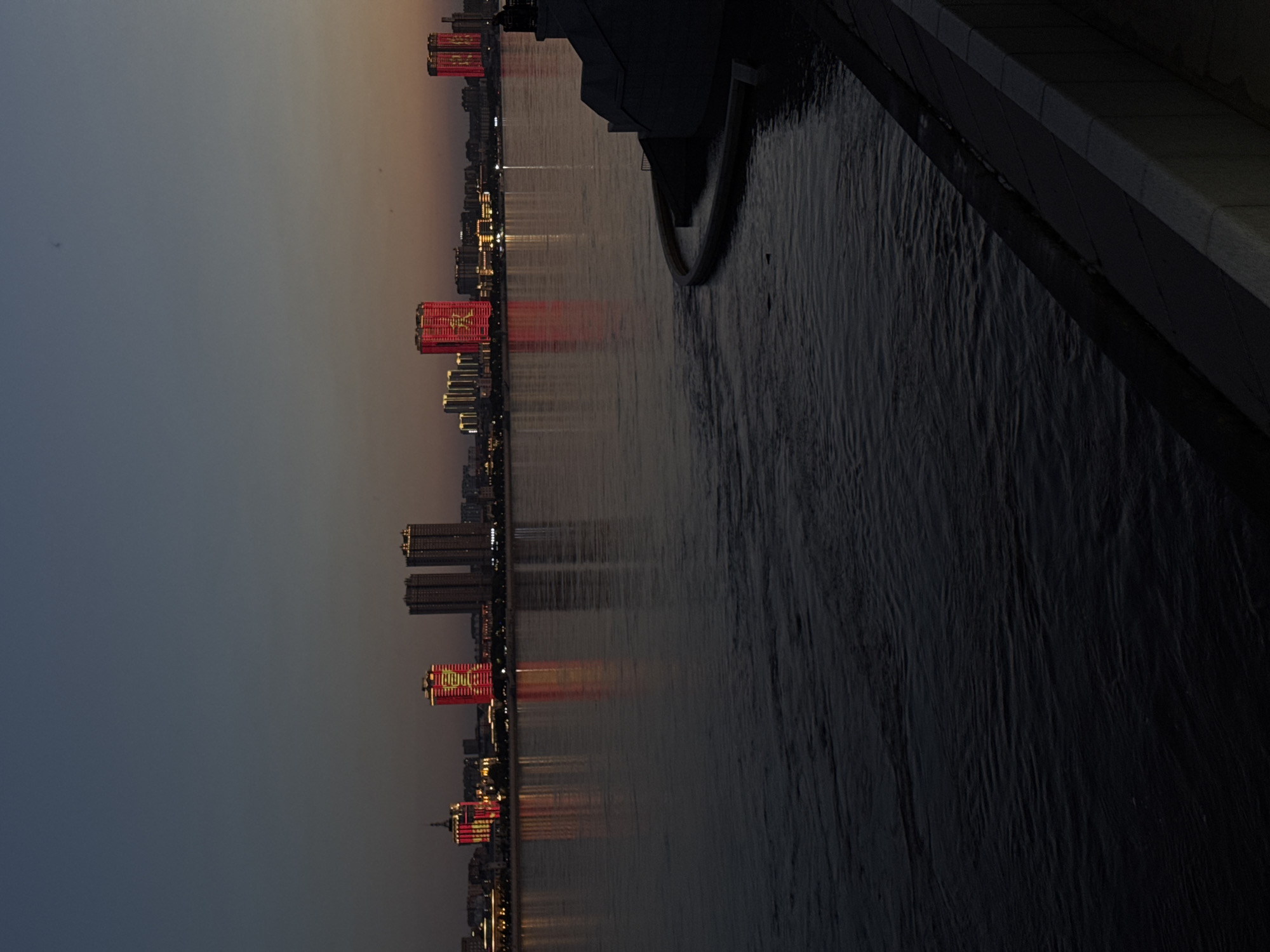 This is a photograph of a cityscape at dusk, likely in Blagoveshchensk, Russia. The image is oriented vertically, with the buildings appearing on the left side and a body of water on the right.
The main subjects are the buildings, which are illuminated by a warm, red light. Several of these buildings display large, red banners with yellow text. One banner prominently shows what appears to be a Chinese or Korean character, while another has a star symbol. These illuminated structures are reflected in the dark, rippling water. In the distance, other buildings are visible with their lights on, creating a sparkling effect.
The time of day is dusk, indicated by the dim, gradient sky transitioning from a light orange near the horizon to a deep blue. The water's surface is textured with ripples, suggesting a slight breeze. The right side of the image shows a dark, paved embankment or walkway bordering the water, with a section of dark, possibly concrete, structure that angles away from the viewer.
The overall mood is one of a bustling city at twilight, with the bright lights contrasting against the fading daylight and dark water. There is no discernible activity of people, only the static presence of the architecture. The visible text on the banners is red with yellow lettering.