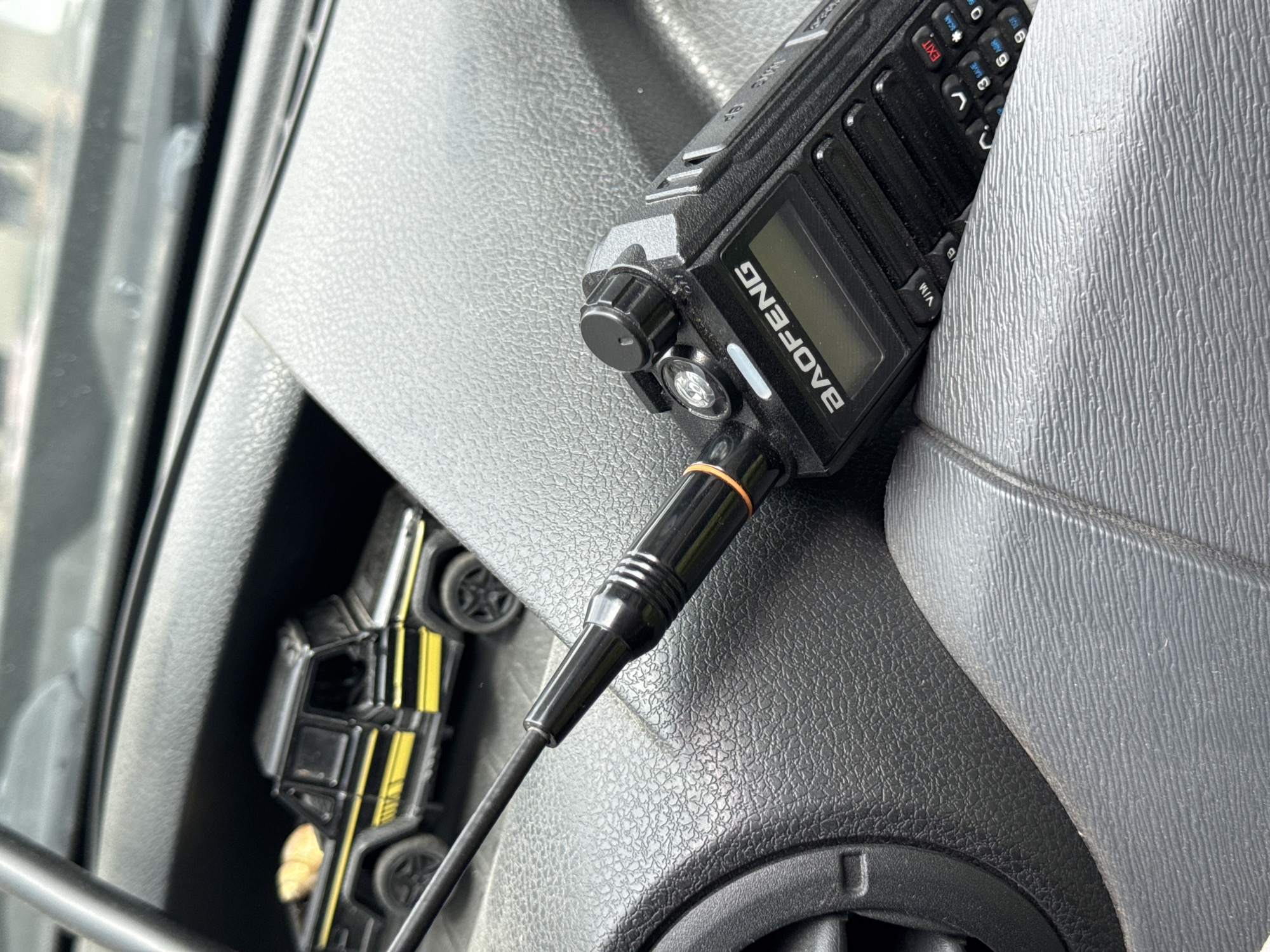 A black Baofeng handheld radio is positioned within the textured gray dashboard of a vehicle. The radio is connected to a black cable. A toy car, featuring black and yellow coloring, is partially visible to the left of the radio. The location cannot be determined from the image.