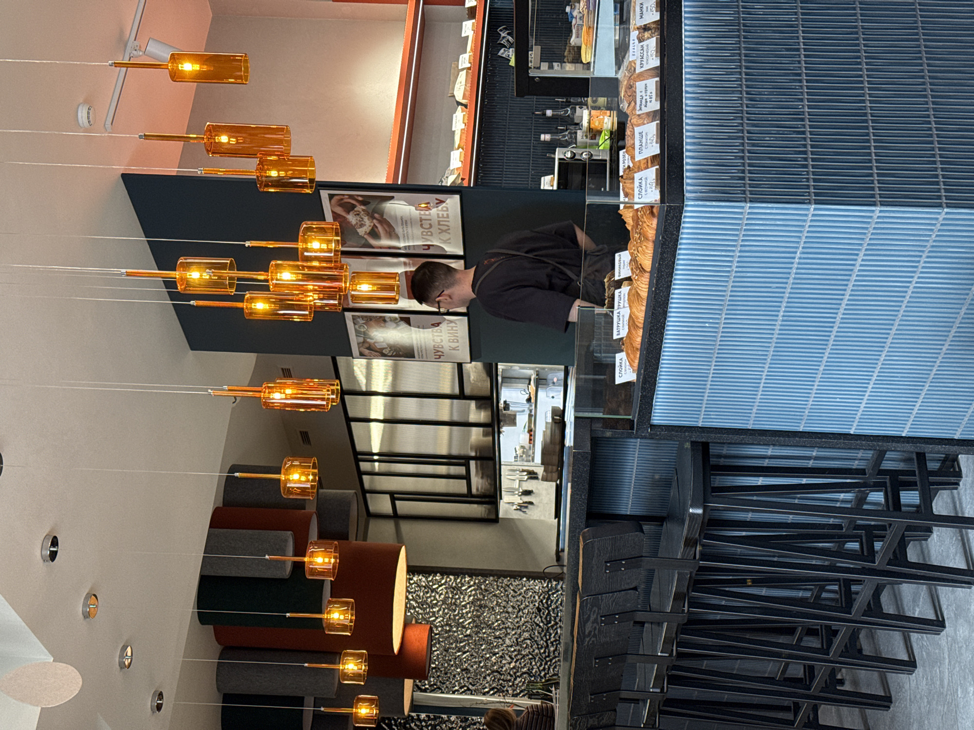 A vertically oriented photograph captures the interior of a shop or bakery in Krasnoyarsk, Russia. A male shop assistant stands behind a glass display case filled with pastries and price tags written in Russian, indicating items like "croissant" and "pastry with cottage cheese." He wears a dark shirt and glasses, his attention directed towards the display.
Above the display, posters bearing Russian text and images adorn the wall. A modern lighting fixture composed of vertically stacked orange glass cylinders descends from the ceiling, illuminating the area. The floor features a textured design, while a stack of black chairs is visible in the foreground, suggesting a seating area for customers. The overall ambiance of the shop is modern and stylish.