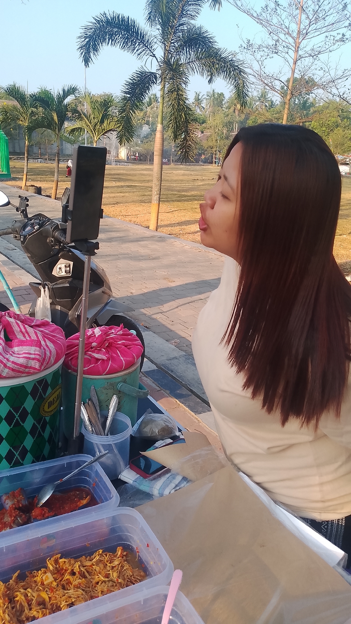 A young woman with long brown hair is captured in a side profile, puckering her lips while looking towards a smartphone mounted on a selfie stick. She is wearing a light-colored shirt. The scene appears to be outdoors, with palm trees and a grassy field visible in the background, suggesting a location like Karanglangu, Indonesia, possibly near a park or public space. In the foreground, there is a mobile food stall with two large containers covered with pink and white striped cloths, and several plastic containers filled with food. A motorcycle is parked nearby, and the ground is paved with patterned tiles. The lighting indicates it is daytime, likely during the morning or late afternoon due to the warm, soft sunlight.