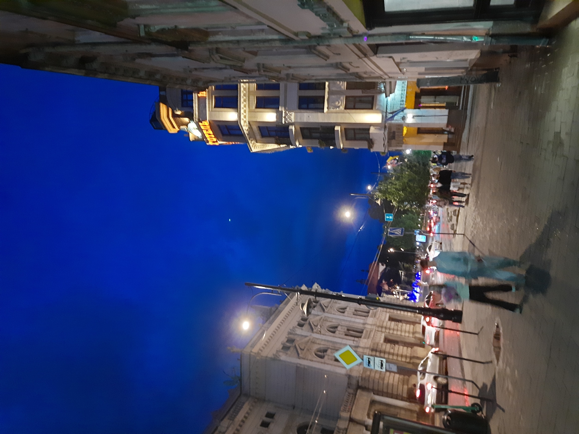 The image shows a street scene in Lviv, Ukraine at dusk. On the left is a large, ornate building with classical architectural features. A yellow diamond-shaped sign with a bus symbol is visible on a pole in front of it. Across the street, a tall, multi-story building with illuminated windows and a prominent "HOTEL" sign in yellow Cyrillic lettering is visible. Two people, a male and female, are walking on the sidewalk in the foreground, the female carrying a bouquet of flowers. Several cars are visible in the street, some with their headlights on. The sky is a deep blue, indicating twilight.