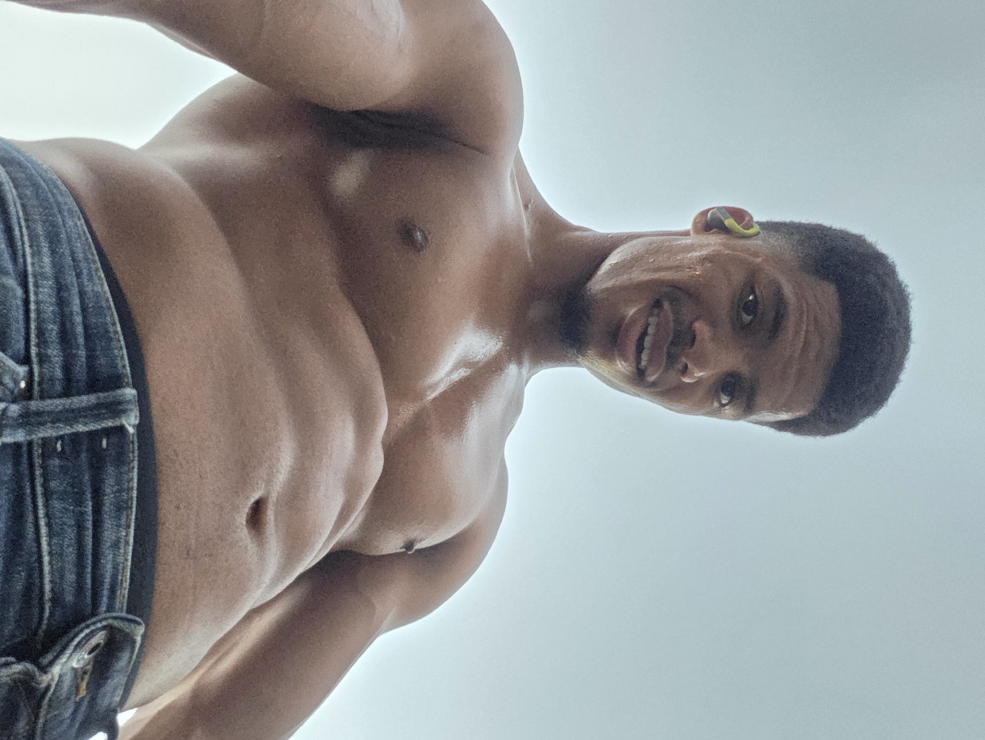 A low-angle, waist-up shot captures a young Black man, shirtless and wearing blue denim jeans, looking directly at the camera with a slight smile. His skin is glistening with sweat, suggesting recent physical activity. He has short, dark, curly hair and is wearing a green and yellow earbud in his left ear. The background is a bright, overcast sky, providing no discernible urban context for Varna, Bulgaria. The image is taken outdoors, and the lighting suggests it is daytime.