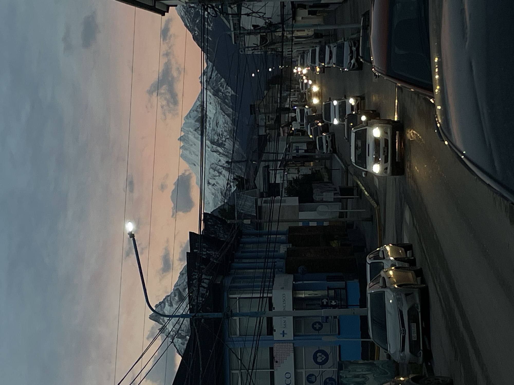 The image depicts a street in Ushuaia, Argentina, with a view of snow-capped mountains in the background. The street is lined with buildings on the left, including a building with a blue facade and signs that partially read "SAN FRANCISCO." Overhead power lines are visible. Cars are present on the street, both parked and driving, with headlights illuminated. The sky displays a gradient of colors, suggesting either sunrise or sunset. The overall scene appears to be a residential or commercial area within the city.