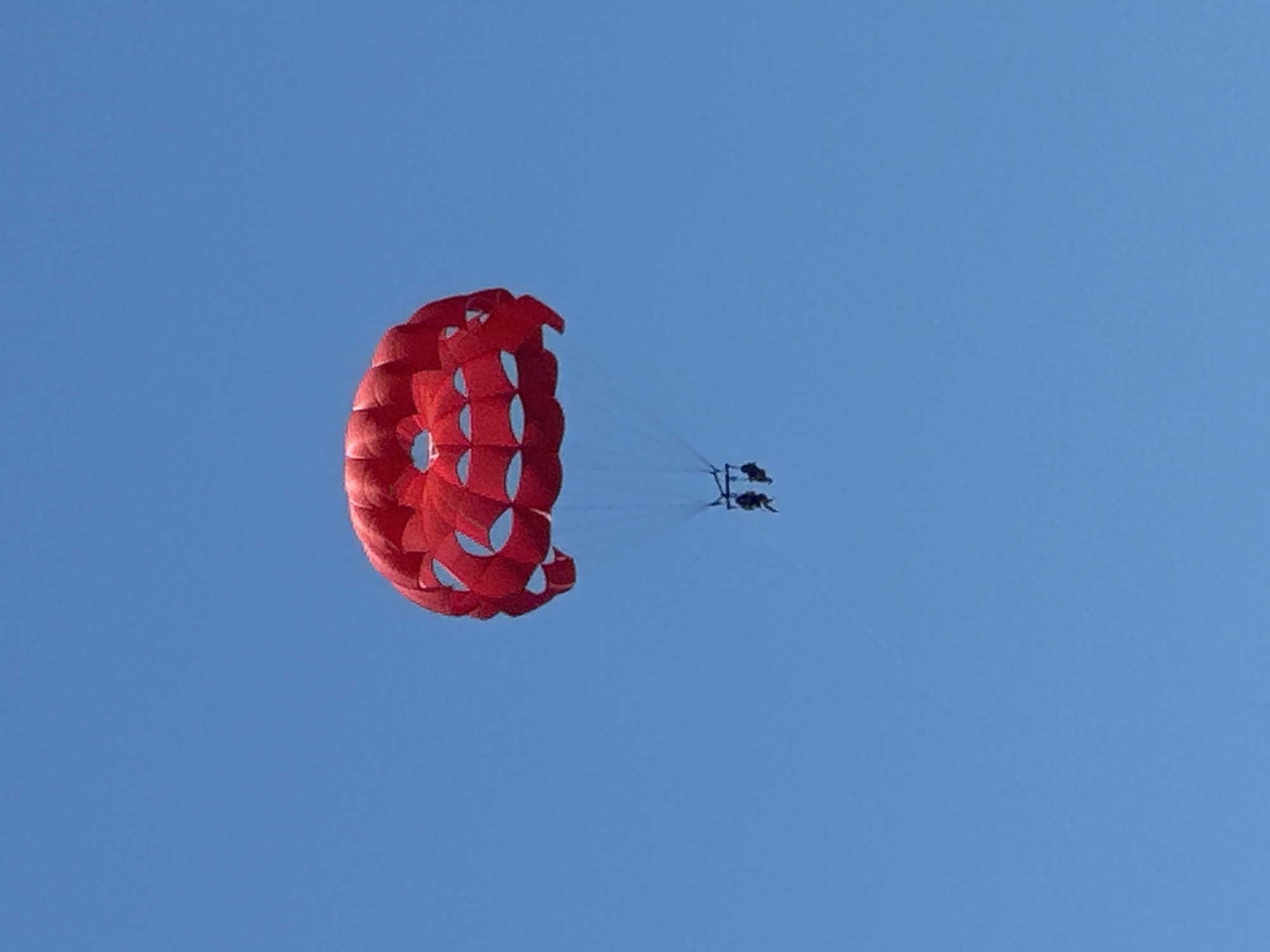 Two individuals are parasailing under a bright red parachute against a clear blue sky. The parachute features a distinct circular pattern with triangular cutouts. The people are suspended below the parachute by ropes, appearing as dark silhouettes against the sky. The scene is consistent with an outdoor recreational activity in a coastal or open area. The location is identified as Boreti, Montenegro, suggesting a seaside resort or a similarly scenic spot.