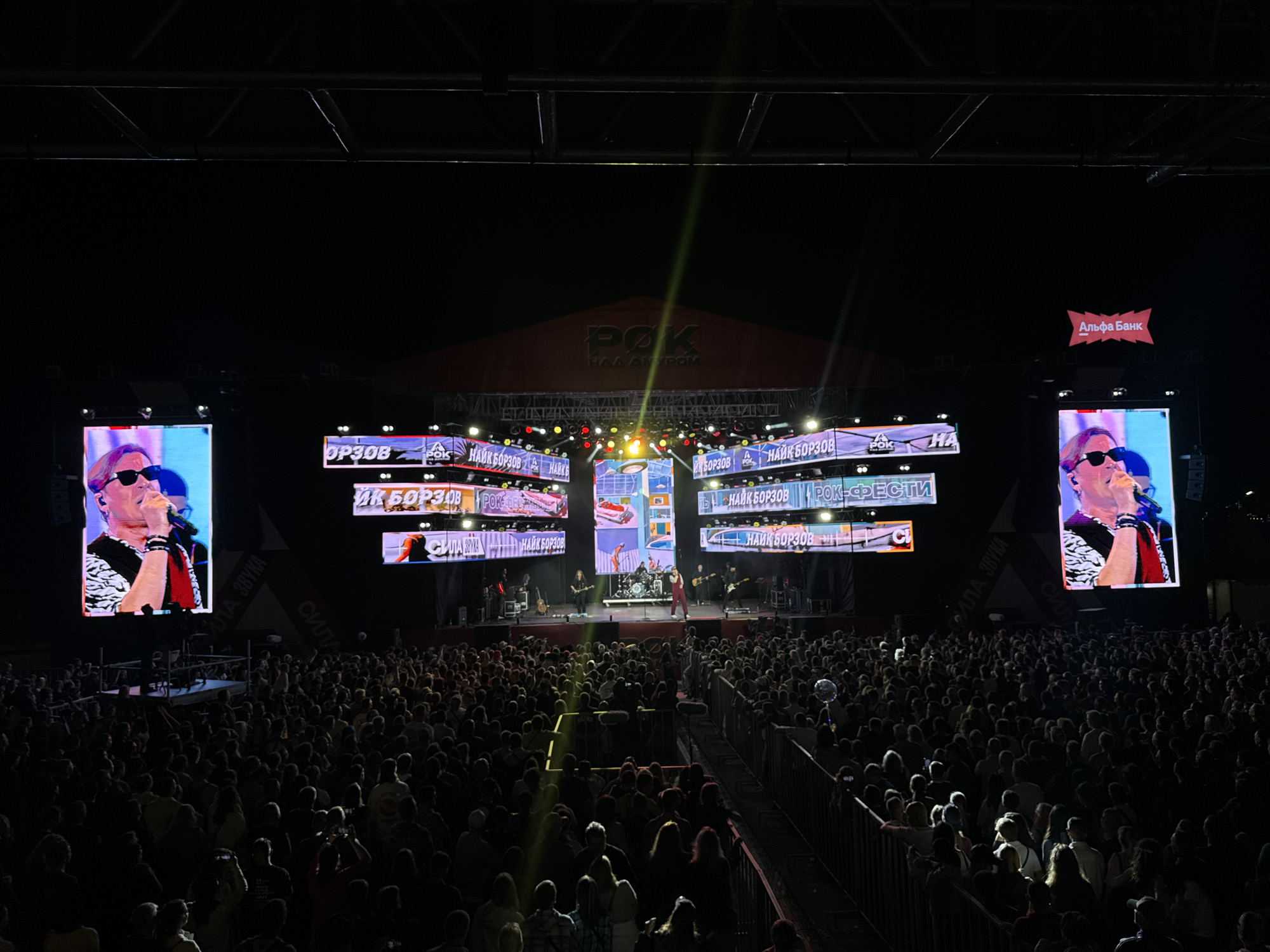 The image depicts a large outdoor concert event in Khabarovsk, Russia. The main focus is a brightly lit stage with multiple large LED screens displaying imagery of performers and event branding. One screen on the left shows a close-up of a male singer in sunglasses and a black and white patterned shirt, holding a microphone. To the right, another large screen shows a similar image of a male singer. The central screens display the text "РОК НА ДВ" (Rock on Amur) and "НАЙК БОРЗОВ" (Naik Borzov), indicating the event is a rock festival featuring the artist Naik Borzov. The stage itself is populated with musicians, including a drummer and guitarists, under theatrical lighting. A large crowd of spectators, numbering in the thousands, is visible in the foreground, gathered in front of the stage. The audience is densely packed, with some individuals holding up phones to record. The atmosphere appears to be energetic, with the concert in full swing. The overall environment is dark, suggesting it is nighttime, with the stage lighting and screen illumination providing the primary light sources. The event appears to be sponsored, with a prominent "Альфа Банк" (Alfa-Bank) logo visible on a banner. The geographical location is identified as Khabarovsk, Russia, based on the text displayed on the screens.