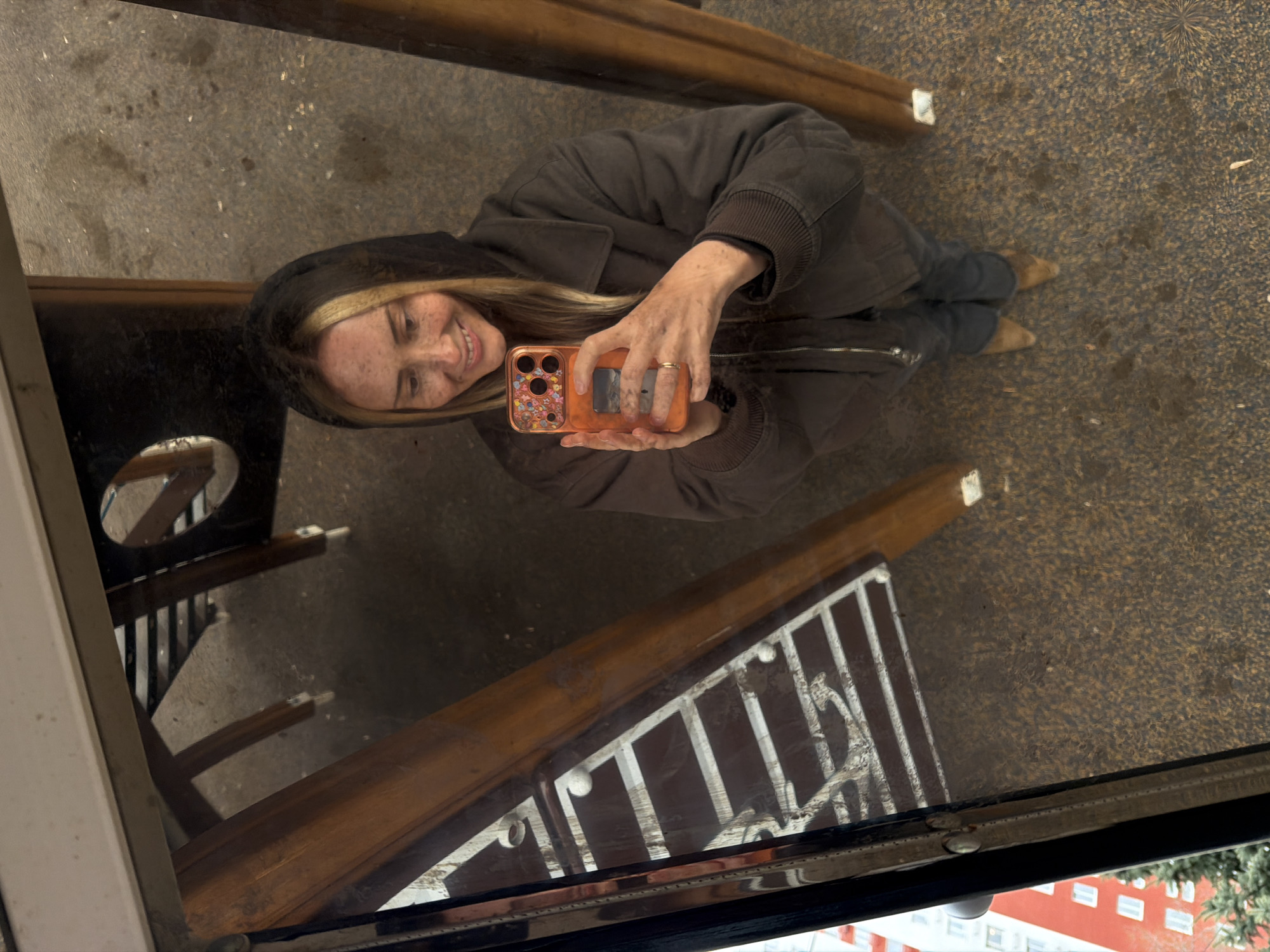 An overhead shot captures a young woman taking a reflection selfie in what appears to be a large, reflective, possibly wet or polished surface, creating an unusual perspective. The woman is smiling, looking down and slightly to her right, with her head tilted. She has blonde hair, visible freckles on her face, and wears a dark grey or brown hooded jacket. Her lower body, dressed in dark trousers and light brown pointed-toe shoes, is also visible.
She holds an orange smartphone with a distinctive multi-colored, circular patterned case in both hands, positioned to capture her reflection. A simple gold-colored ring is on her right-hand ring finger.
The reflective surface on which she appears to be "lying" has a textured, granular appearance, resembling asphalt or a paved ground. Through the reflection, elements of an urban environment are visible: a red building with multiple white-framed windows, green foliage (likely trees), and decorative metal railings or fencing, suggesting a public space in a city like Moscow, Russia, as specified by the location context. The lighting suggests daytime, possibly overcast, given the soft, diffused light and lack of harsh shadows. No legible text is visible in the image.