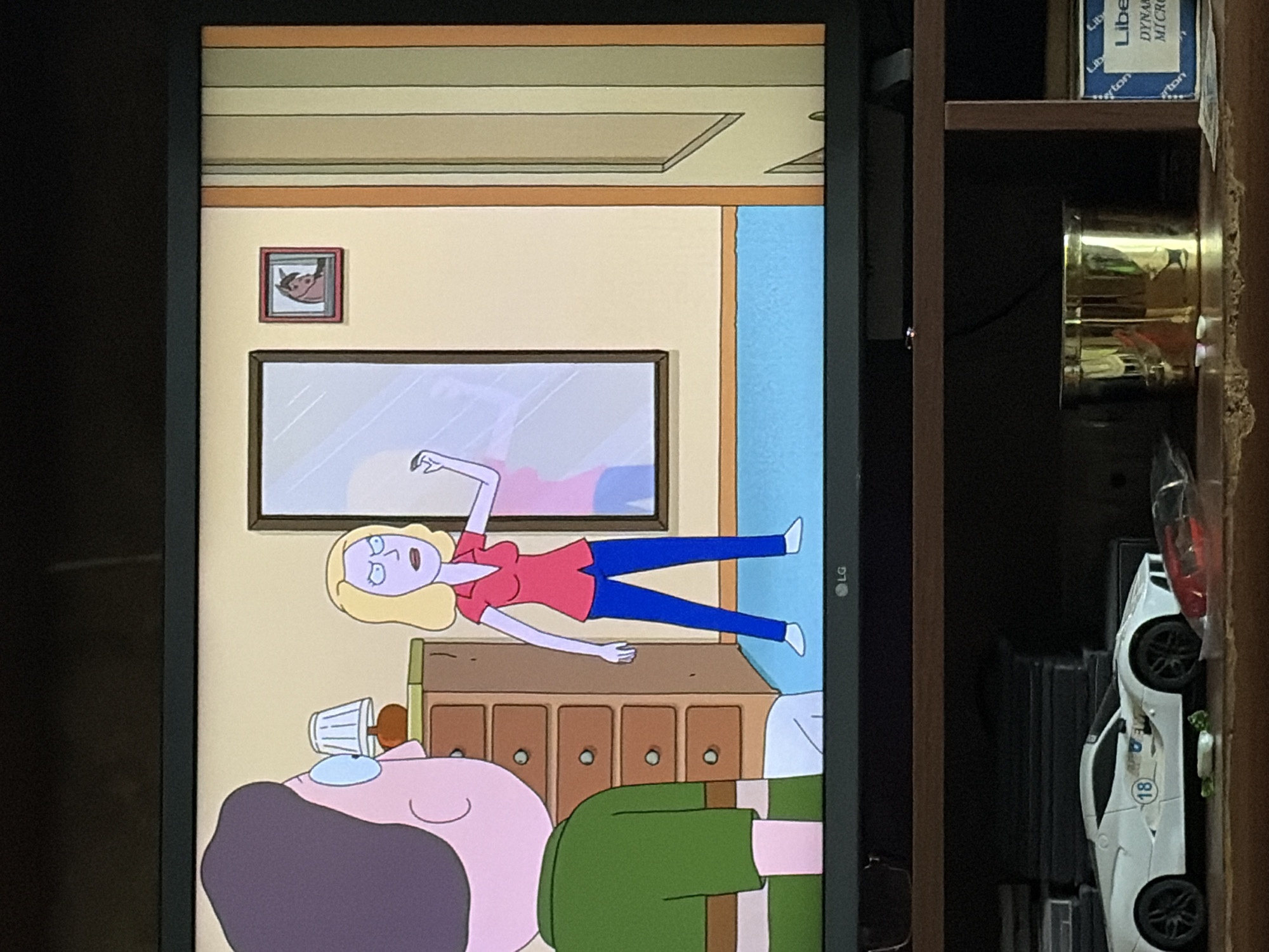 A television screen displays a scene from an animated show. In the foreground, to the left, a character with purple hair and glasses, wearing a green shirt, is seen from the back. To the right of this character, a blonde woman with a purple complexion, wearing a pink shirt and blue pants, stands facing a mirror. Her expression is stern, and she appears to be gesturing with her right hand. Behind her, on the wall, is a small framed picture of a horse's head. The environment is a room with light yellow walls, a wooden dresser with multiple drawers, and a blue floor. Below the television screen, a shelf holds various items including a white toy sports car with the number 18 on its side, a golden cylindrical object, and a blue box with white text.