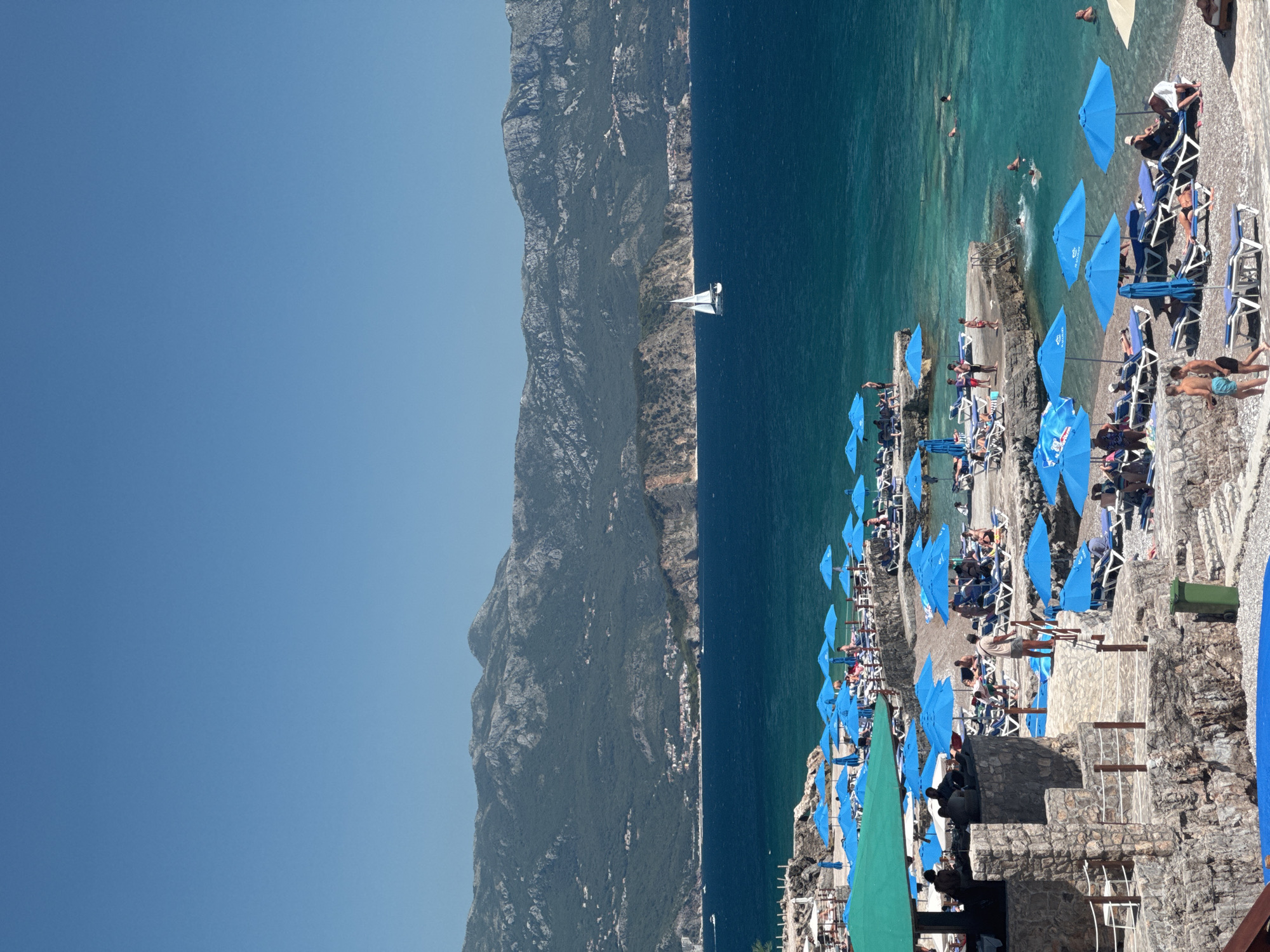 The image depicts a vibrant coastal scene in the city of Kubasi, Montenegro, under a clear, bright blue sky. In the background, a range of rugged, forested mountains with visible rock formations stretches across the horizon, with scattered settlements discernible on their lower slopes. The deep blue sea occupies the middle ground, transitioning to a clearer turquoise closer to the shore. A white sailboat with its sails unfurled is visible on the open water.
The foreground features a multi-tiered beach area constructed with natural stone walls and pathways. Numerous light blue beach umbrellas provide shade over rows of white lounge chairs with blue cushions, occupied by individuals. People are seen relaxing, sunbathing, and walking on the beach terraces. On the left, a prominent stone structure with a green canopy, likely a beach bar or restaurant, hosts several patrons. Further right and closer to the water, multiple individuals are swimming or wading in the clear, shallow sea. A green trash bin is visible on one of the lower stone terraces.