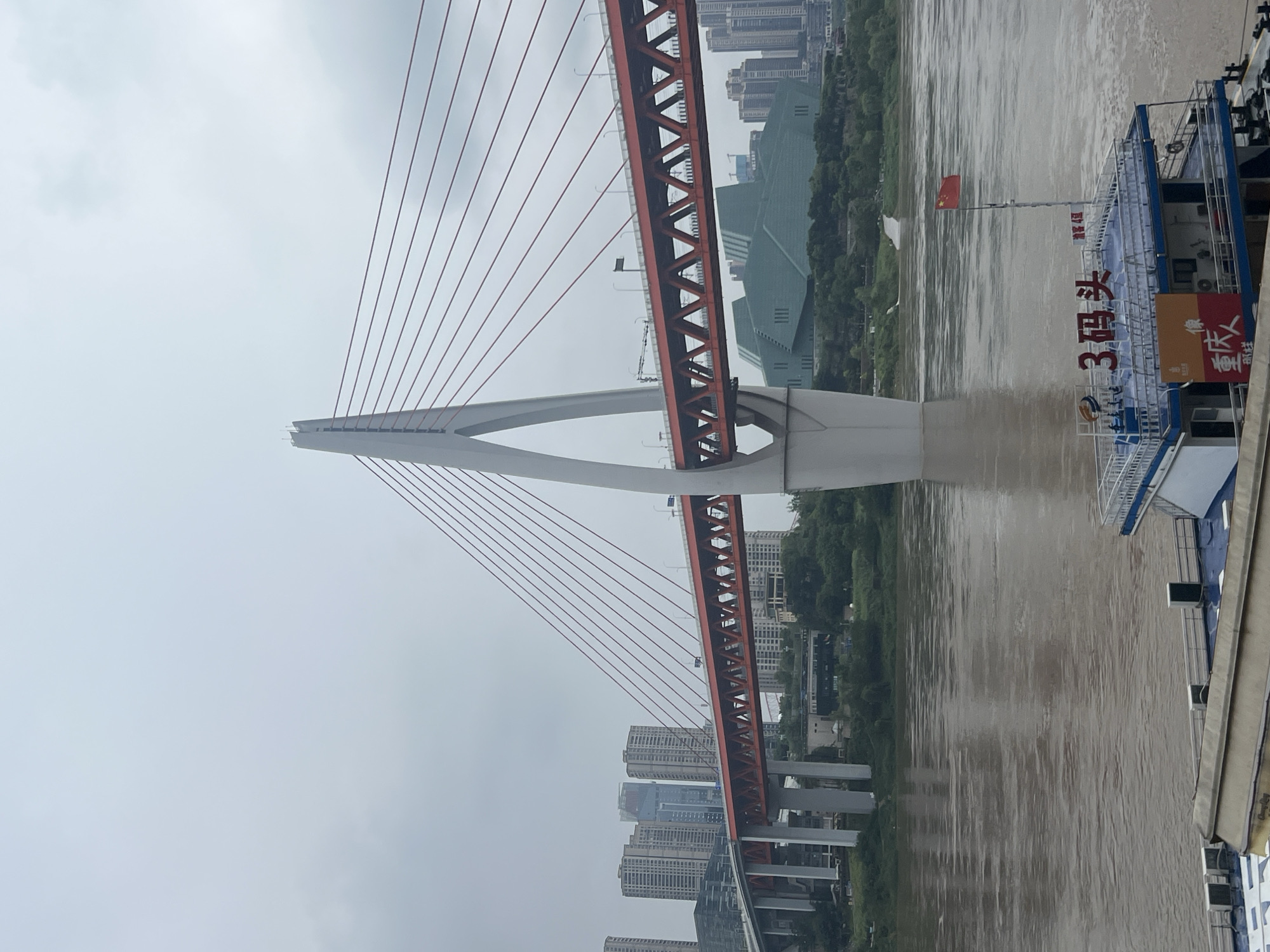 The image shows the Chaotianmen Bridge in Yuzhong District, Chongqing, China. The bridge is a cable-stayed bridge with a distinctive white pylon and red cables. It spans a wide, muddy river, likely the Yangtze River, which is a significant geographical feature of the city.
On the bridge, traffic can be seen, indicating its function as a transportation artery. In the background, a cluster of modern high-rise buildings forms the city's skyline, characteristic of a major urban center. A large, green, geometric building is also visible near the riverbank, possibly a cultural or administrative facility.
In the foreground, part of a boat is visible, with a Chinese flag flying from a pole. The boat has text in Chinese characters, including "3号码头" which translates to "Pier 3" and other markings that are unreadable. The boat appears to be docked or stationary on the water. The overall atmosphere of the image is somewhat overcast, suggesting a cloudy day. The scene captures a blend of modern infrastructure and natural elements, typical of Chongqing.