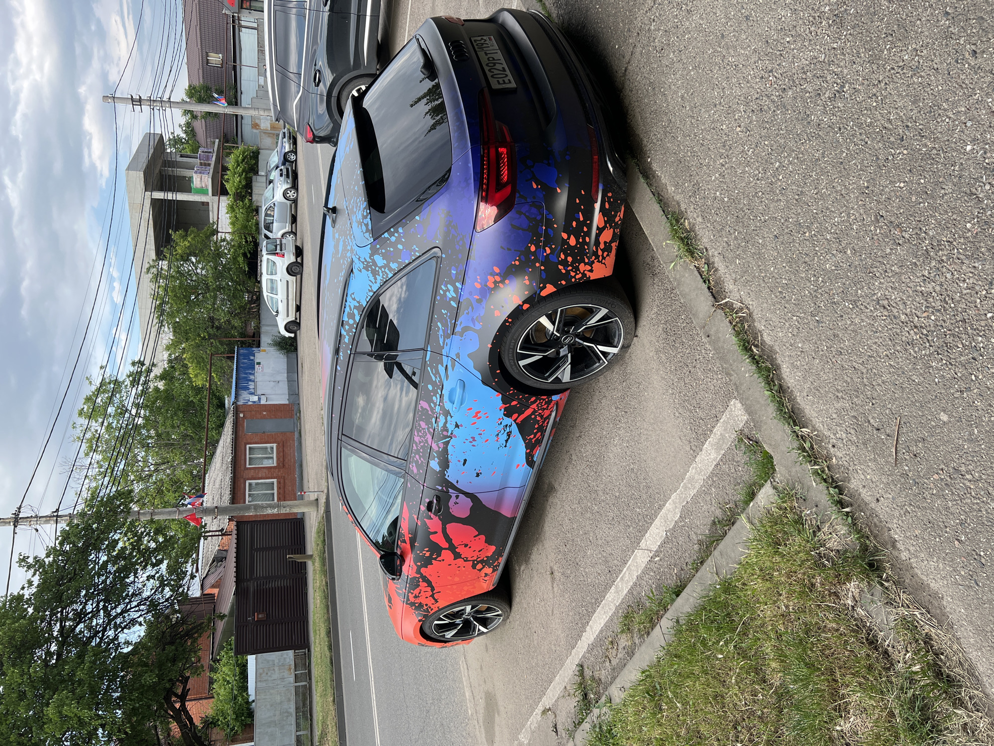 A dark-colored Audi A4 Avant station wagon with a vibrant, multi-colored splatter wrap (predominantly red, blue, and purple) is parked on an asphalt surface, angled slightly towards the right of the frame. The car is positioned in what appears to be a parking area next to a curb with green grass. The background shows a street with other vehicles, including a dark SUV to the right and several cars parked further down the street. Buildings, including a brick house with a metal fence and a partially constructed concrete building, are visible in the distance, along with trees and utility poles. The sky is overcast with clouds. The license plate of the Audi is visible and reads "E029PT 193". The image was taken from a slightly elevated viewpoint, looking down at the car.