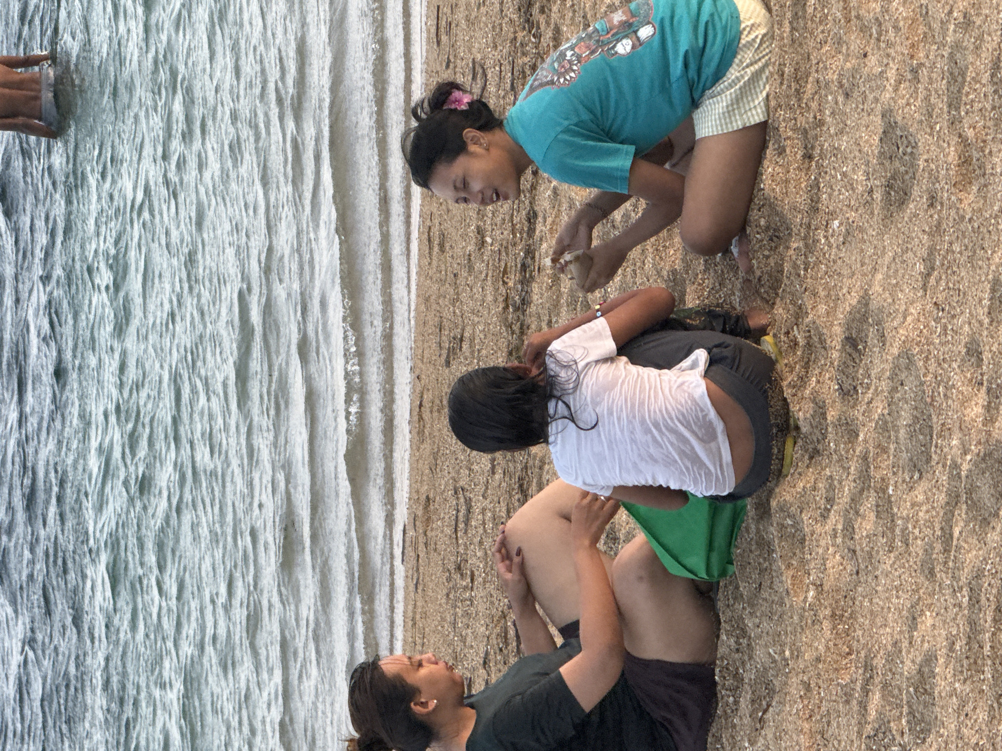 Three young women are sitting on a sandy beach, facing the ocean. A man is partially visible in the water in the background. The woman on the left is wearing a black t-shirt and dark shorts. The woman in the middle is wearing a white t-shirt and dark pants. The woman on the right is wearing a teal t-shirt and light-colored shorts, and has a pink flower in her hair. The ocean water is choppy, with small waves. The sand on the beach appears to be composed of small shells and pebbles. The scene is likely Kuta Beach in Denpasar, Indonesia, known for its lively atmosphere and wide sandy shores.