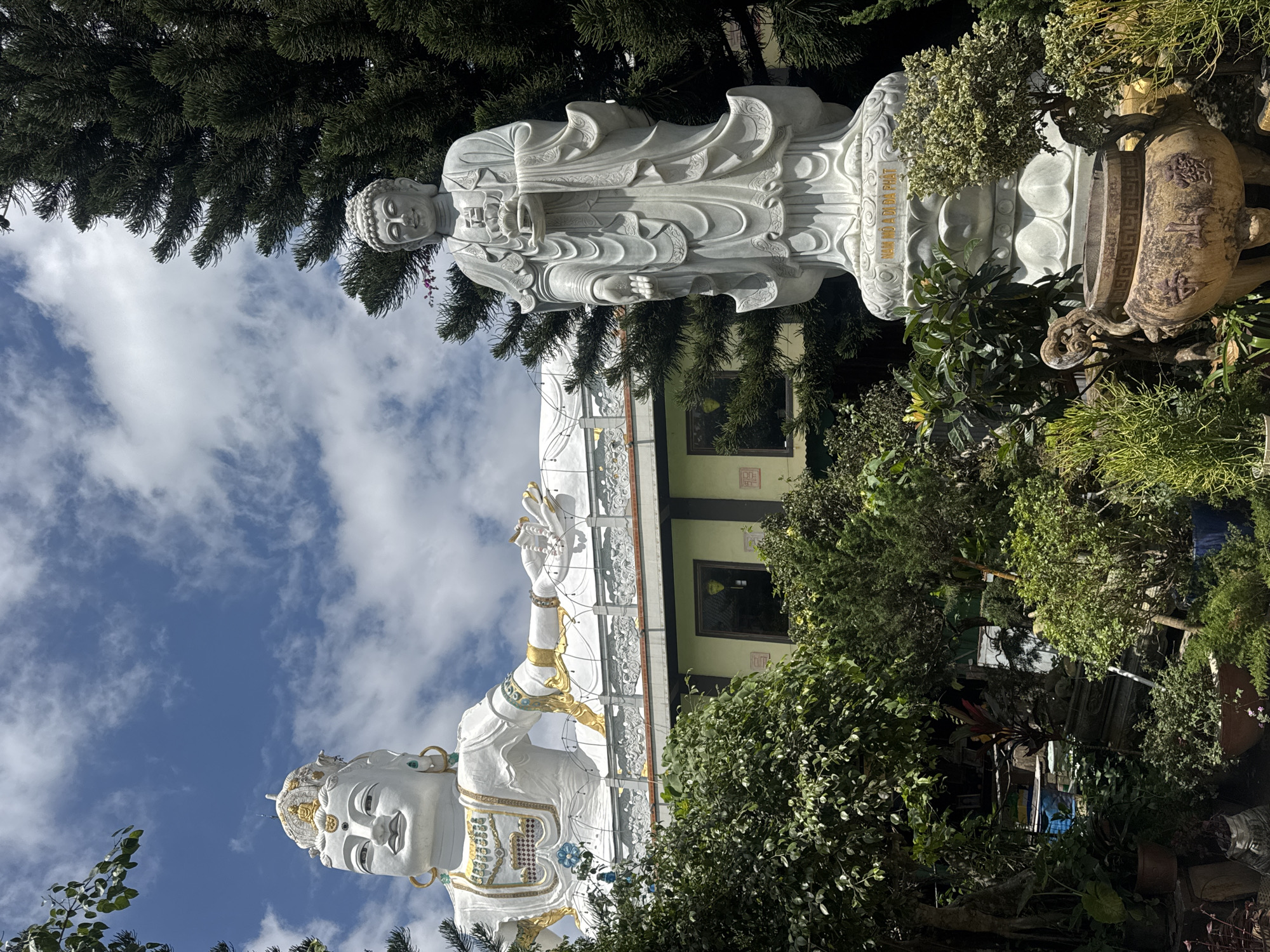The image displays two large statues in front of a building, set against a backdrop of a blue sky with scattered clouds and lush green trees. In the foreground, a large, intricately carved stone urn is partially visible, adorned with Chinese characters. To its right, a tall, white marble statue of a standing figure, possibly a deity or a revered person, holds an object in its cupped hands. The figure is dressed in flowing robes, and its base is inscribed with Vietnamese text. Behind this statue, a massive white statue of a reclining figure, likely a Buddhist deity, dominates the upper portion of the image. This colossal figure, adorned with gold accents and jewelry, rests on a white structure that appears to be part of a building. The building itself has a light-colored facade with dark-trimmed windows. The surrounding area is densely planted with various types of green foliage, including trees and smaller bushes, suggesting a garden or a landscaped temple complex. The overall impression is one of serene grandeur and spiritual significance. This scene is located at the Linh Phuoc Pagoda in Da Lat, Vietnam, a popular tourist destination known for its unique architecture and impressive Buddhist sculptures.