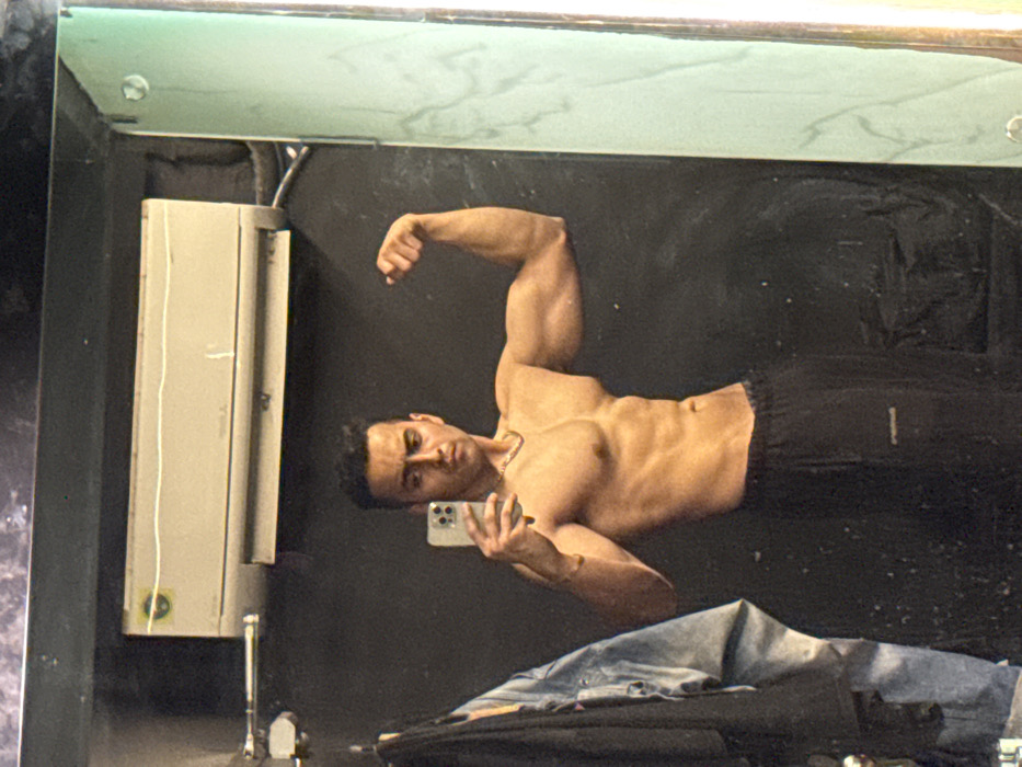 A shirtless man flexes in a bathroom mirror, holding a phone in his left hand to capture the moment. He's wearing dark athletic shorts. The background includes a grey industrial-style electrical box mounted on the wall. A discarded pair of jeans rests on a surface below the mirror. The scene is slightly rotated clockwise.
