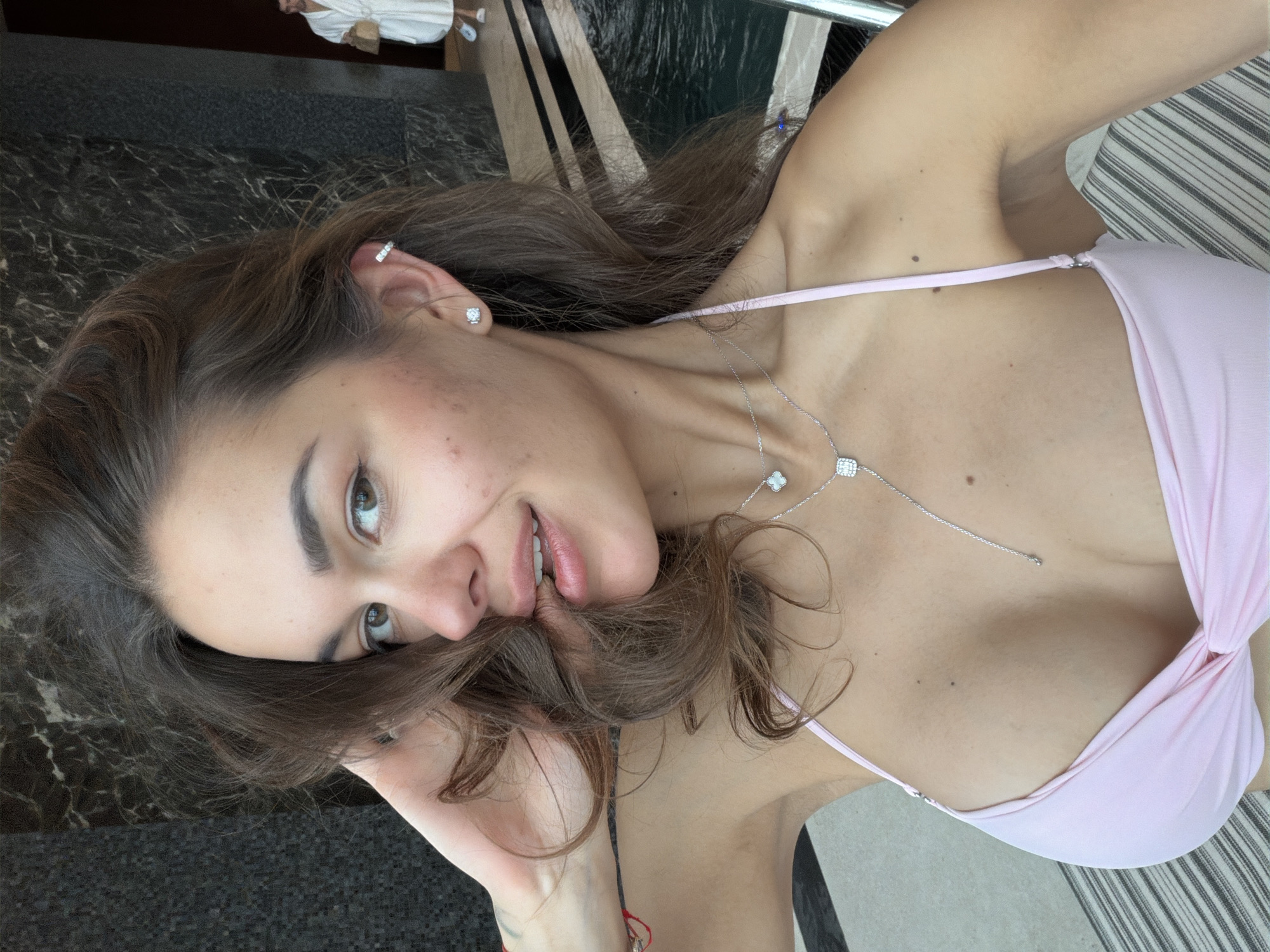 A selfie of a young woman with long brown hair wearing a pink bikini and silver necklace. She is partially obscuring her face with her hair. She has fair skin, brown eyes, and pierced ears. The background includes a dark marble floor, a stripe pattern, and a glimpse of a person in a white robe. The location is not discernable from the image.