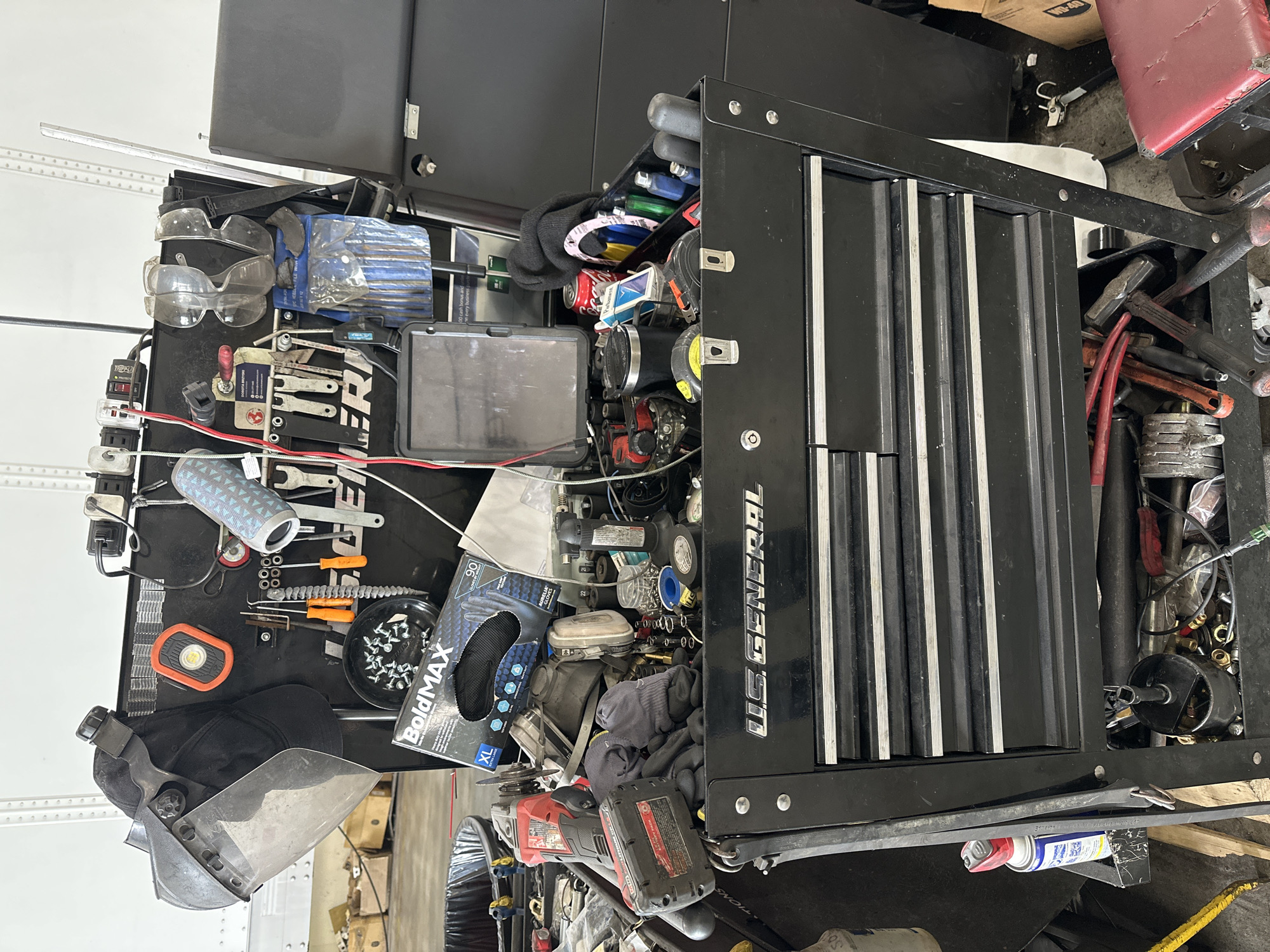 An outdoor shot of a US General tool chest in Burr Ridge, Illinois, showcases a collection of tools and safety equipment. A black baseball cap with a face shield attached is hanging from the top left of the chest, next to an orange magnetic work light. Below these, a box of BoldMAX XL gloves rests on top of a pile of other gloves. The chest itself is filled with various tools, including screwdrivers, pliers, and what appears to be a power drill with a battery pack. On the right side of the chest, a pair of safety glasses hangs above a blue tool pouch. A can of Coca-Cola and a pack of cigarettes are also visible on top of the chest, near a roll of paper towels. The chest is situated in what looks like a workshop or garage, with industrial shelving and boxes visible in the background.
