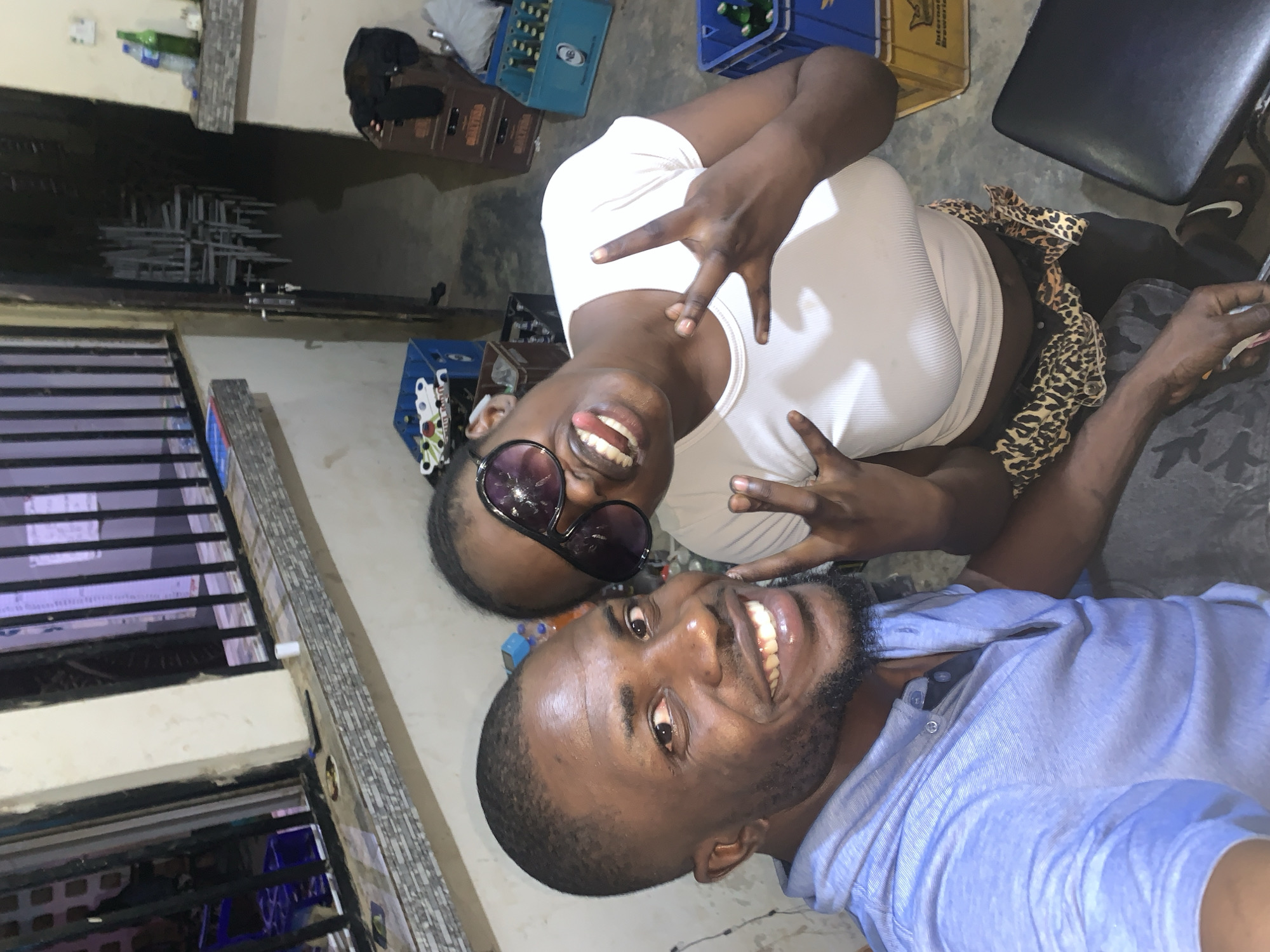 Two people are posing for a selfie, with a male in the foreground wearing a light blue shirt and smiling, and a female in the background wearing sunglasses and a white shirt, making a hand gesture. They are indoors, with various objects in the background including crates of drinks, seating, and a barred window. The location is in the city of New Stadium, Nigeria.