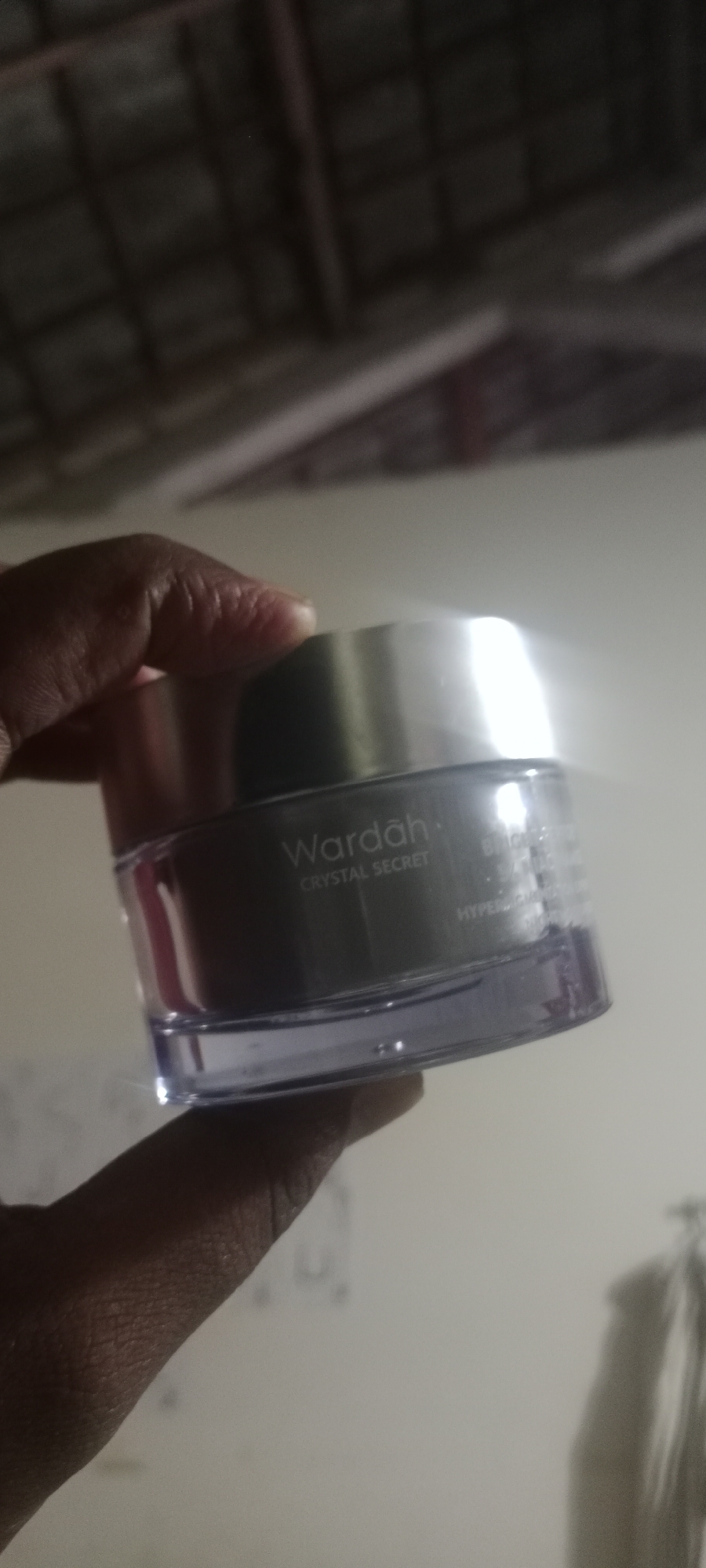 A close-up shot shows a hand holding a jar of Wardah Crystal Secret cosmetic product. The jar is made of clear plastic with a silver lid. The label on the jar reads "Wardah Crystal Secret" and some other text that is not fully legible. The background is out of focus, but appears to be an indoor setting with a light-colored wall. The lighting is bright, causing some glare on the lid of the jar. The image is taken from a slightly elevated angle. The city of Soko, Indonesia is not discernible from this image.
