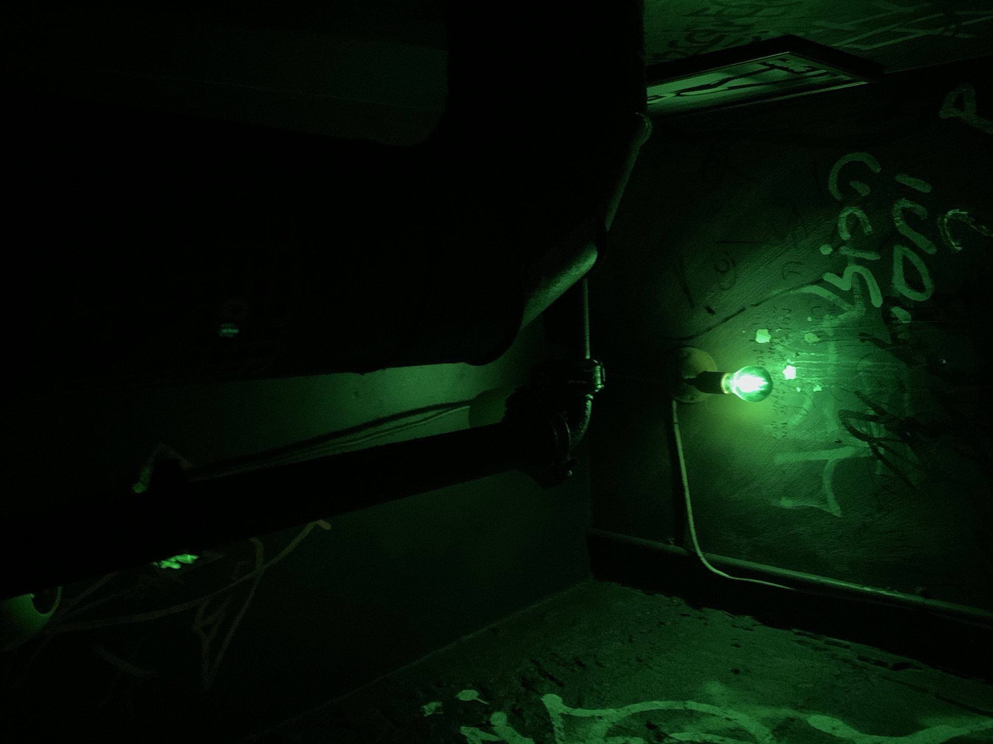 The image depicts an interior scene illuminated by a single green lightbulb. The environment appears to be a dimly lit, possibly abandoned or industrial space, with a rough, textured wall covered in graffiti. A pipe runs along the wall, and electrical wiring is visible near the ceiling. The graffiti on the wall is in a dark, rough texture, with lighter green spray paint applied over it. Some of the graffiti appears to be in Hebrew script, including visible letters that could spell out phrases or words. The overall impression is one of a gritty, urban setting in Tel-Aviv, Israel, characterized by its subdued lighting and weathered surfaces.