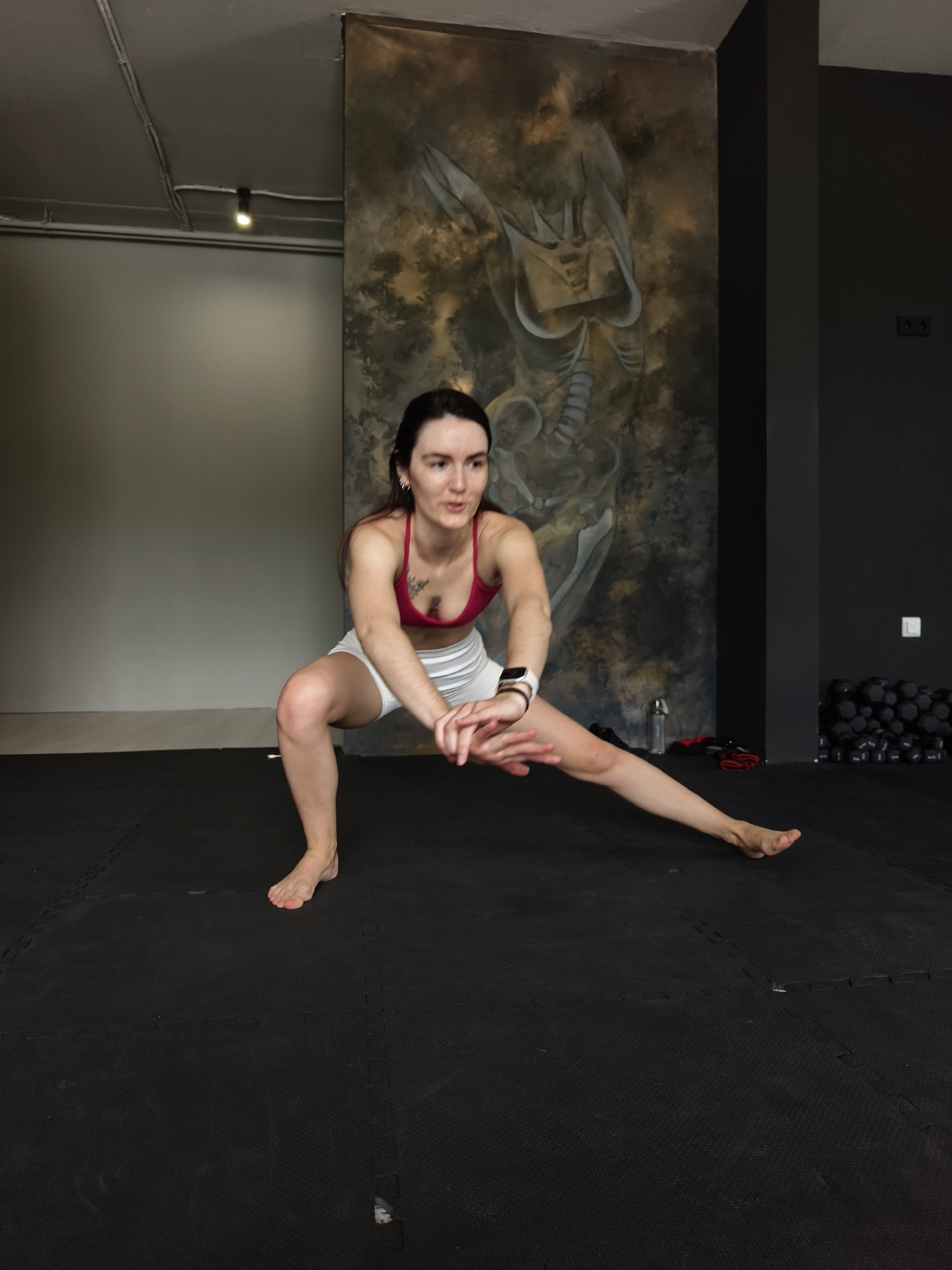 A woman is depicted performing a side lunge exercise in an indoor gym or fitness studio in Tyumen, Russia. The subject has dark hair, fair skin, and is wearing a red sports bra, white shorts, and a smartwatch on her left wrist. She is barefoot and has a tattoo on her upper chest. Her left leg is bent, with her body lowered, while her right leg is extended straight out to the side. Her hands are clasped in front of her, and she has a focused expression.
The setting features black interlocking foam mats covering the floor. The walls vary, with a plain light gray on the left and a darker gray/black on the right. A prominent, large mural on the dark wall behind the woman displays abstract, skeletal, or anatomical figures in earthy, muted tones. In the background on the right, a stack of black dumbbells and a water bottle are visible, indicating a workout area. Overhead lighting illuminates the space. No discernible text is visible in the image.