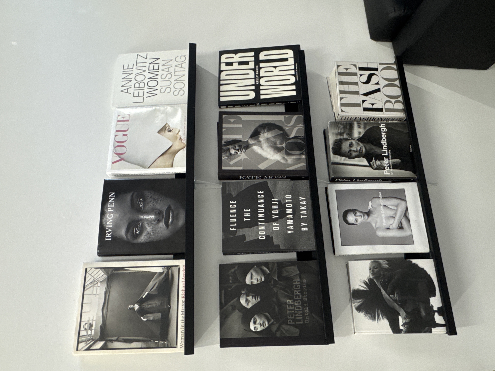 The image shows three stacks of books arranged on a surface, possibly a bookshelf or the floor. The left stack contains books titled "Woman in the Mirror Richard Avedon," "IRVING PENN," "VOGUE," and "ANNIE LEIBOVITZ WOMEN SUSAN SONTAG." The middle stack displays books titled "PETER LINDBERGH Untold Stories," "FLUENCE THE CONTINUANCE OF YOHJI YAMAMOTO BY TAKAY," "KATE MOSS," and "HUNDER WORLD KELLY KLEIN." The right stack showcases books titled "Patrick Demarchelier," "Peter Lindbergh Onn Fashion Photography," and "THE FASE BOOL." The environment is a room with bright, neutral-toned walls. There is a dark-colored object, possibly furniture, in the upper right corner of the image. The location and presence of individuals are not provided in the image.
