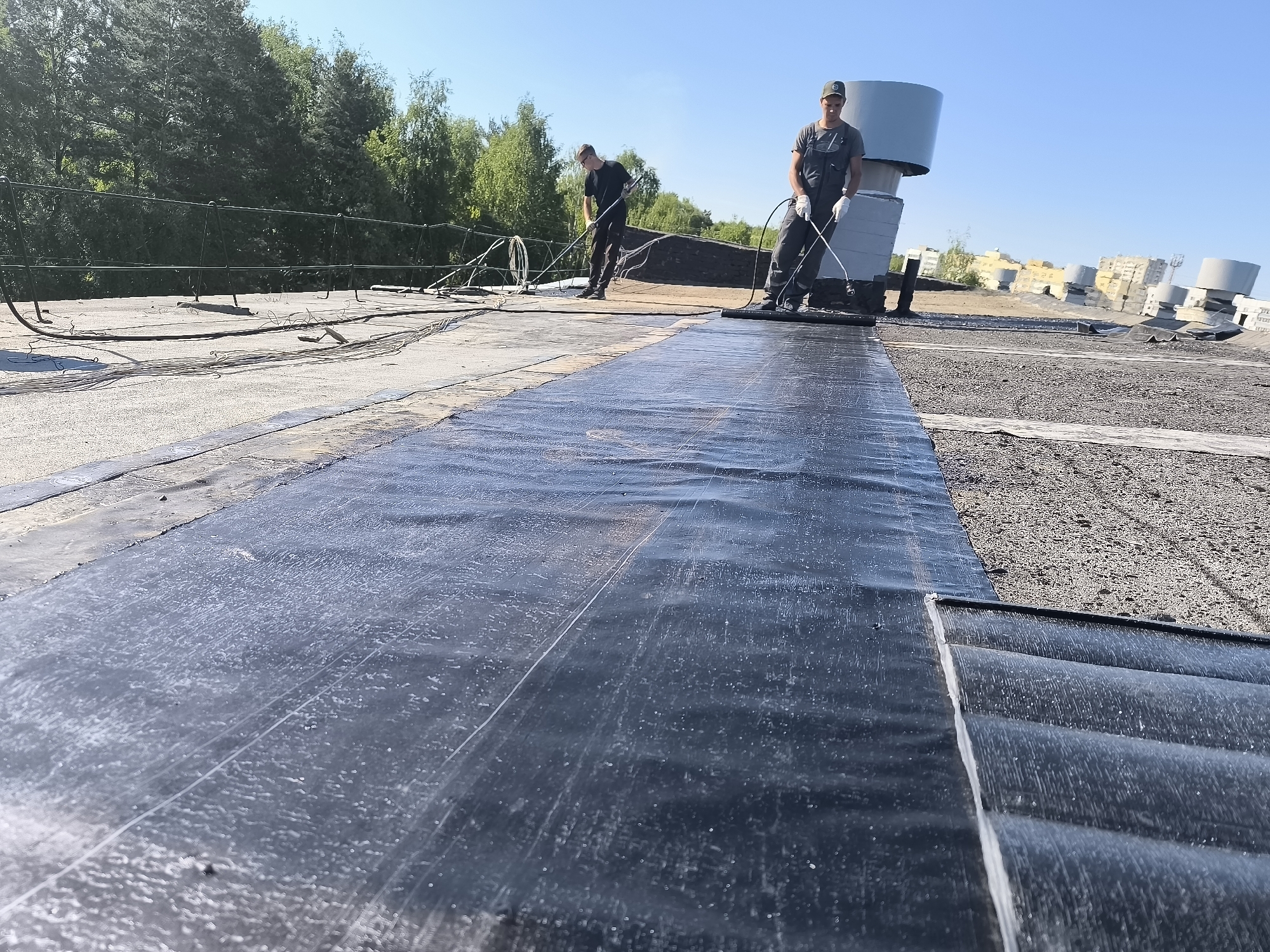The image depicts two workers applying a black, bituminous waterproofing membrane to a flat roof in an unidentified urban area. One worker is seen near the center of the frame, facing right, wearing a grey jumpsuit, work gloves, and a baseball cap. He holds a tool with a long handle and a flat, spreading end, presumably to press the membrane firmly onto the roof surface. The other worker is further away, facing left, dressed in dark clothing. He also holds a tool with a long handle, suggesting he is involved in the same application process.
In the foreground is a newly laid strip of the black membrane, glistening as if recently applied. The area behind the workers displays older roofing materials, including gravel and existing layers of waterproofing. Several cylindrical rooftop structures, possibly ventilation or exhaust units, are visible in the background. A wire fence runs along the edge of the roof in the background on the left, with trees visible behind it. The weather appears clear, with a blue sky above. The location within the city is unknown.