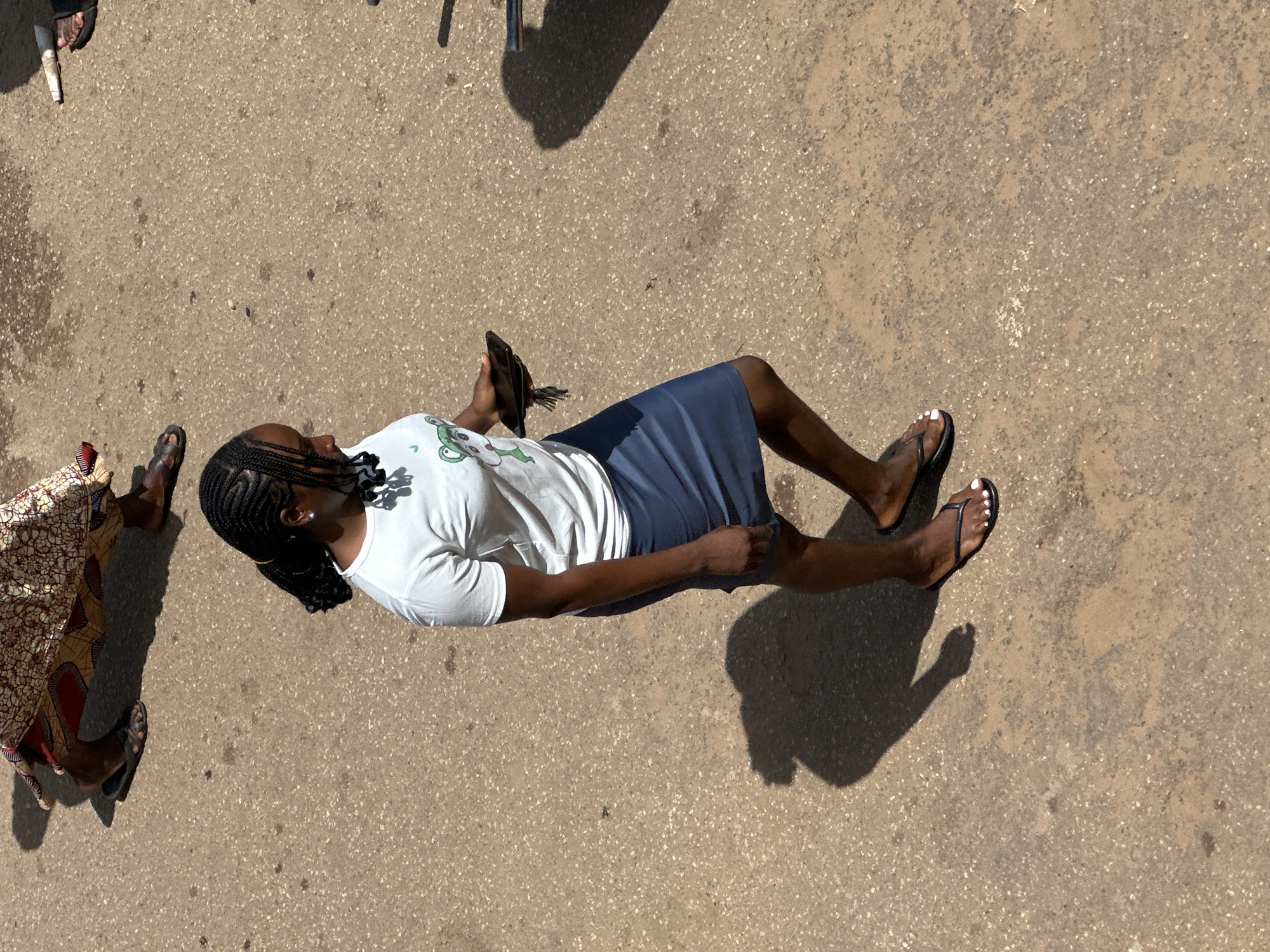 In Bukuru, Nigeria, a young woman with braided hair, wearing a white t-shirt and a blue skirt, walks on a paved surface. She is holding a dark-colored mobile phone in her right hand. Her feet are adorned with black flip-flops. To the upper left of the frame, another person's legs are visible, wearing a patterned cloth garment and sandals. The ground is a textured pavement, casting shadows from the individuals.