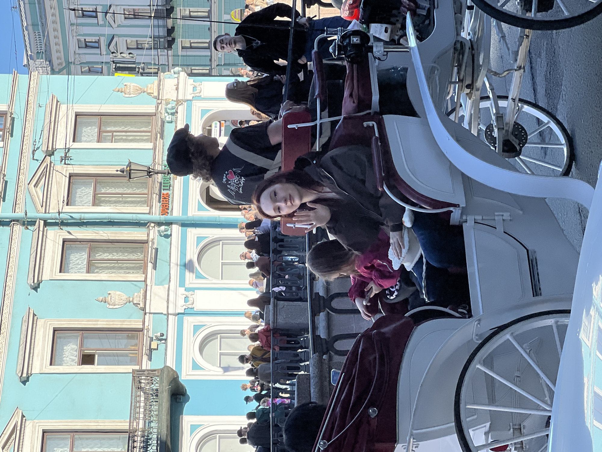 The image shows a group of people riding in horse-drawn carriages near the Winter Canal in Saint Petersburg, Russia. The light blue building is the Winter Palace, visible behind the carriages and people lining the canal embankment. One of the passengers, a young woman with dark hair, is looking directly at the camera. A crowd of people is visible on the embankment, separated from the street by a railing.