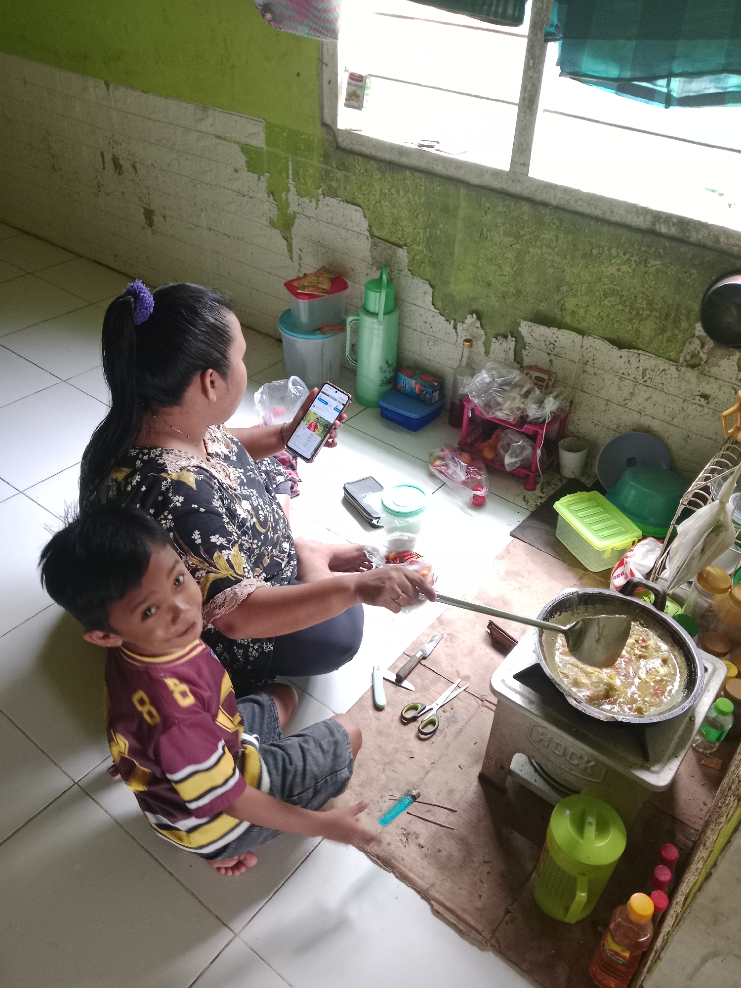 The image depicts an indoor scene, likely a kitchen area, within a humble dwelling in Lakea Satu, Indonesia. A woman, with her dark hair pulled back by a purple scrunchie, is seated on the floor. She wears a dark, floral-patterned top and dark pants. She is holding a smartphone in her right hand, displaying an image of two people. To her left sits a young boy, his gaze directed towards the camera. He is wearing a maroon and yellow-striped jersey with the number 8 prominently displayed. His legs are crossed, and he wears dark shorts.
In the background, a simple stove with a pot of food being stirred by a spatula is visible. Various kitchen items are scattered around, including containers, bottles, a pair of scissors, and a knife. The walls are painted a peeling light green and white, indicating wear and tear. A window with translucent green coverings offers a glimpse of the outside. The floor is tiled in a neutral color. The overall impression is one of everyday life in a modest Indonesian household.
