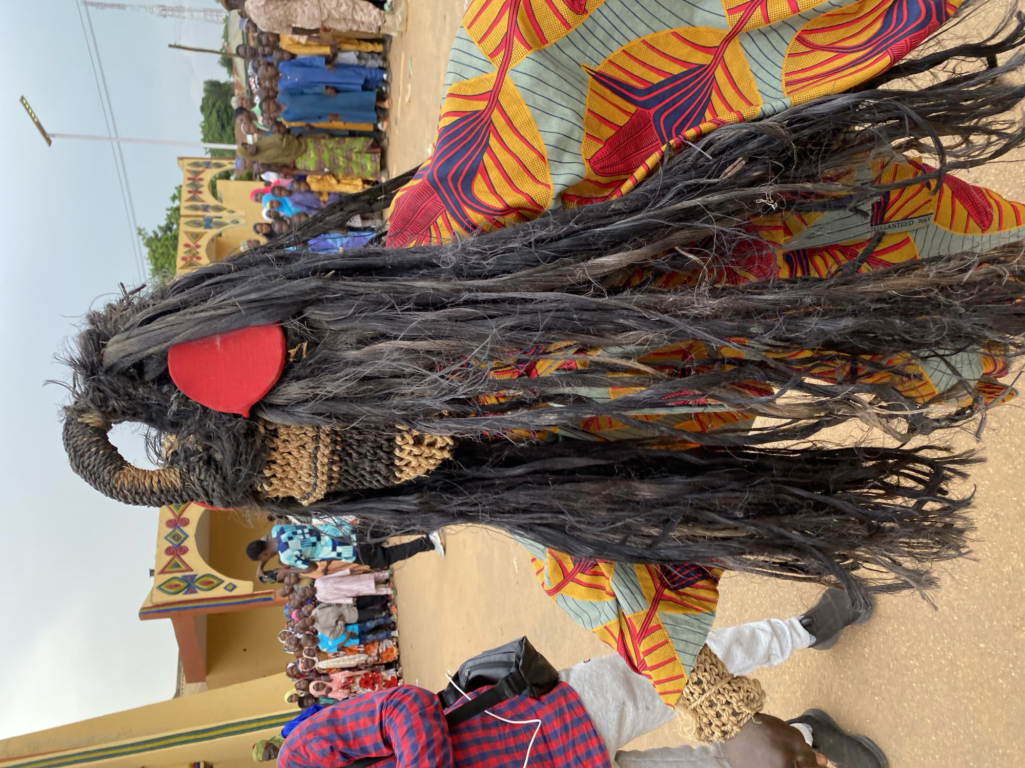 The image shows a person wearing a traditional mask and costume, likely part of a cultural event in Kafanchan A, Nigeria. The mask is characterized by dark, fibrous material resembling hair, a woven section around the face, and a red heart-shaped attachment. The person wearing the mask is also adorned with colorful patterned fabric. The person is wearing footwear. In the background, a crowd of people can be observed near a building with decorated arches and painted walls. A solar light pole is visible.