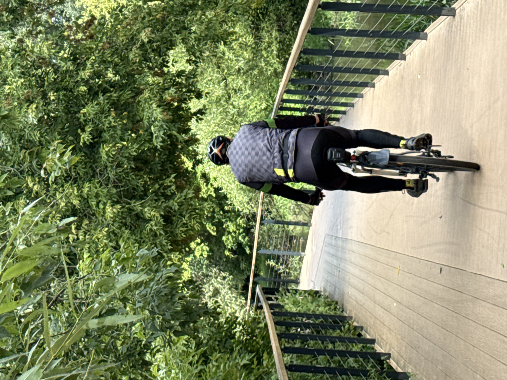 The image shows a person cycling on a wooden boardwalk surrounded by lush green trees and vegetation. The boardwalk has metal railings on both sides. The cyclist is wearing cycling attire, including a helmet, a patterned cycling jersey with green accents, black cycling tights, and cycling shoes. The cyclist is facing away from the camera, moving forward on the boardwalk. The scene appears to be a park or a nature trail within an urban setting, possibly during daytime. There is no discernible evidence to suggest the location is Moscow, Russia.