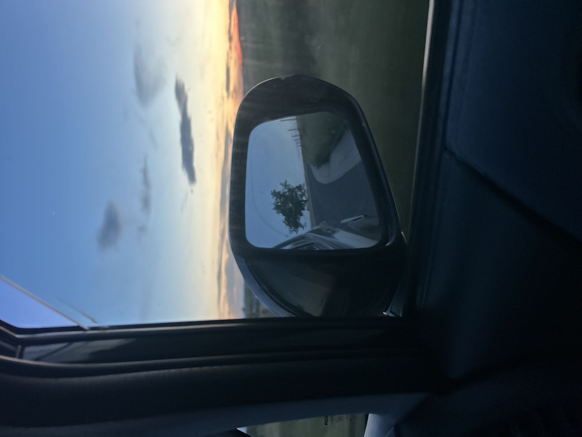 The view from inside a car looking out at the side mirror. The side mirror reflects a road, a tree and another vehicle. The sky and a strip of sunset are visible through the car window. The car's interior is visible, including the door frame and a portion of the dashboard. The location is unknown and there is no evidence to determine that it is in Blatnitsa, Bulgaria. No people are visible.