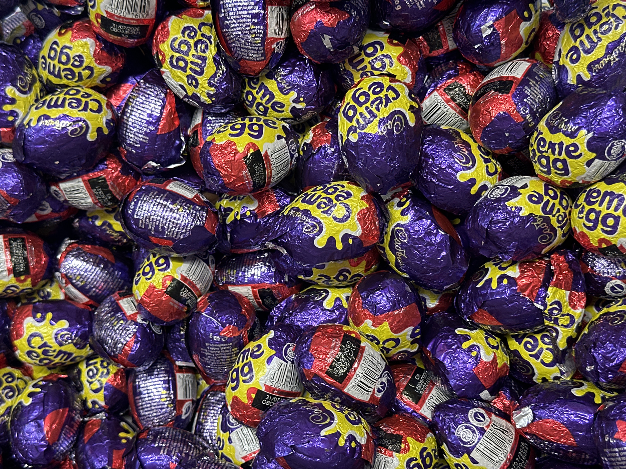 The image depicts a dense pile of individually wrapped Cadbury Creme Eggs. The wrappers are predominantly purple, red, and yellow, with the words "Creme Egg" prominently displayed. Some wrappers also show printed details like barcodes and expiry dates. The focus is tight, creating a close-up view where individual eggs are indistinguishable. The environment appears to be an indoor setting. The location, Wealden, United Kingdom, does not appear in the image, and there is no visual indicator of this.