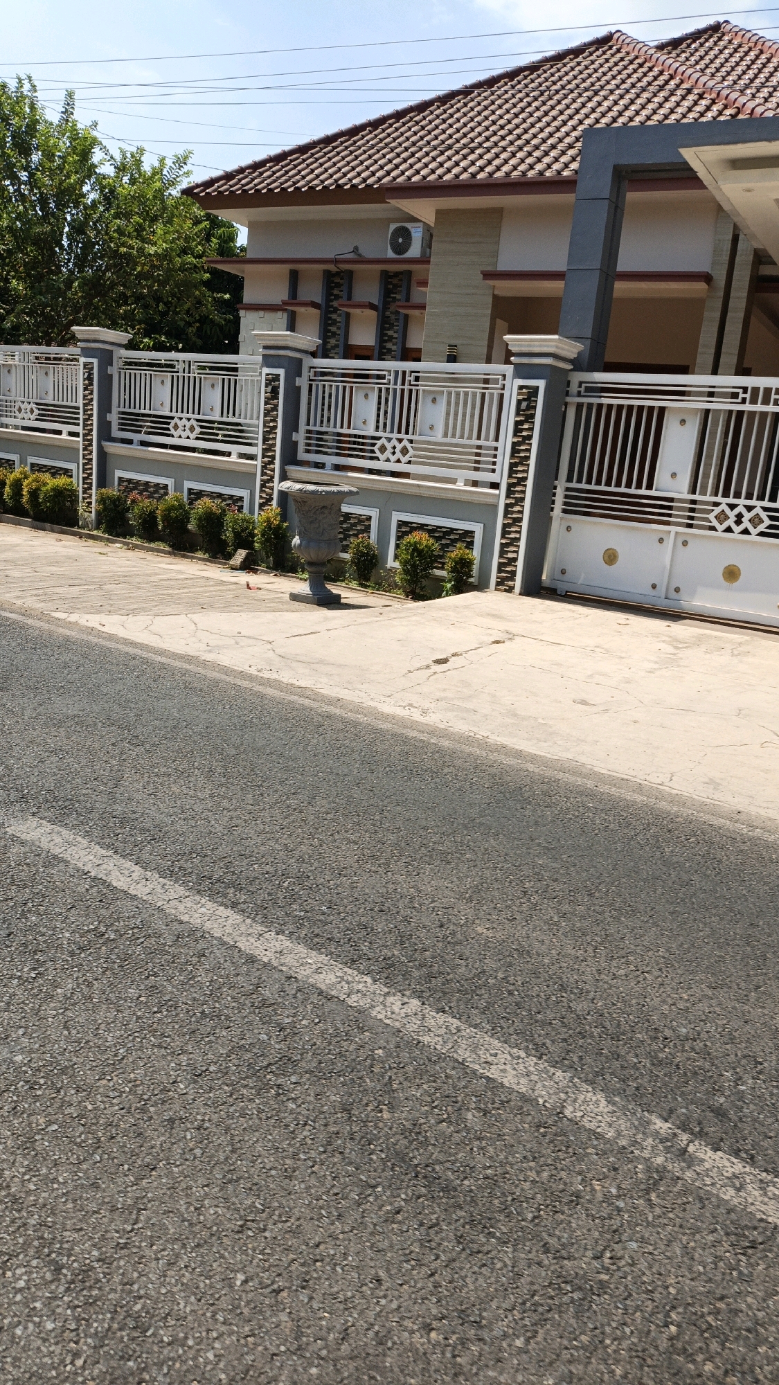 The image depicts a residential property with a white and gray fence and a matching gate. The house, visible behind the fence, has a brown tiled roof and cream-colored walls. An air conditioning unit is mounted on the exterior wall. Small green bushes are planted along the front of the property, and a large decorative pot stands near the gate. The foreground shows a paved road with a white line marking. The scene appears to be in a sunny, outdoor environment.