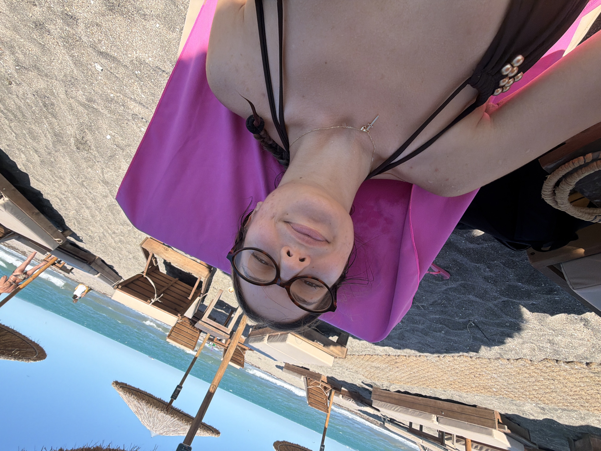 The image shows a selfie taken by a woman lying on a pink towel on a sandy beach. She is wearing glasses and a dark bikini. In the background, there are beach chairs, umbrellas, and the sea. The image is angled, with the woman's face close to the camera. The setting appears to be a beach resort. The image does not provide specific details to pinpoint a location within the city of Burgas, Bulgaria.