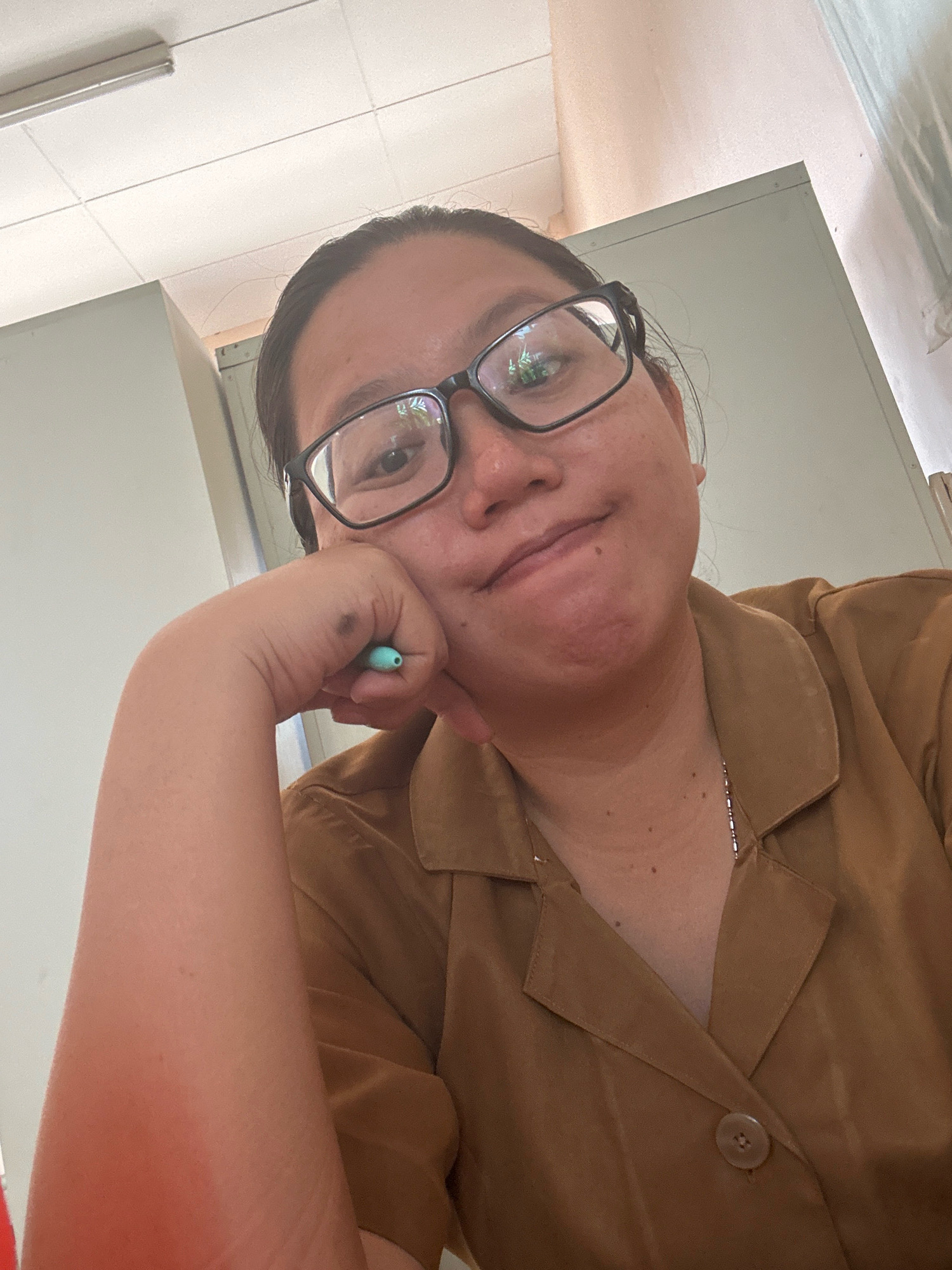 A young woman with dark hair and glasses is resting her chin on her hand, holding a light blue pen. She is wearing a brown collared shirt. The background appears to be an indoor setting with white walls and ceiling. The image is a selfie taken from a slightly low angle. The location is not discernible from the image itself.