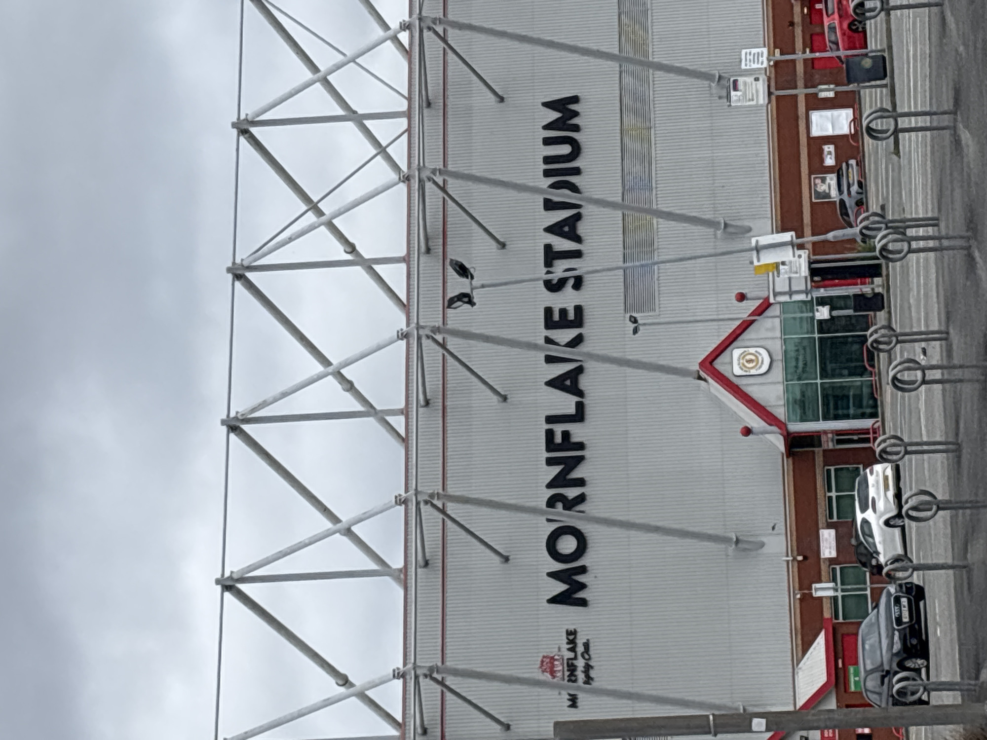 The image shows a view of Mornflake Stadium, located in Crewe, United Kingdom. The stadium facade is a large, ribbed white structure with the words "MORNFLAKE STADIUM" prominently displayed in black lettering. To the left of the stadium, there is a logo featuring a red crest with a bull. Below the logo, smaller text reads "Mornflake - Mighty Oats". The stadium is supported by a complex network of grey metal struts and beams, suggesting a large, modern construction.
In the foreground, there are several cars parked in what appears to be a parking lot or street. A black Audi is on the left, followed by a white SUV, and partially visible grey and red cars further down. Bicycle racks are also visible in the foreground, indicating public access. The sky is overcast, suggesting a cloudy day. The architectural style of the lower part of the stadium, with its red rooflines and glass entrances, suggests a fan zone or hospitality area.