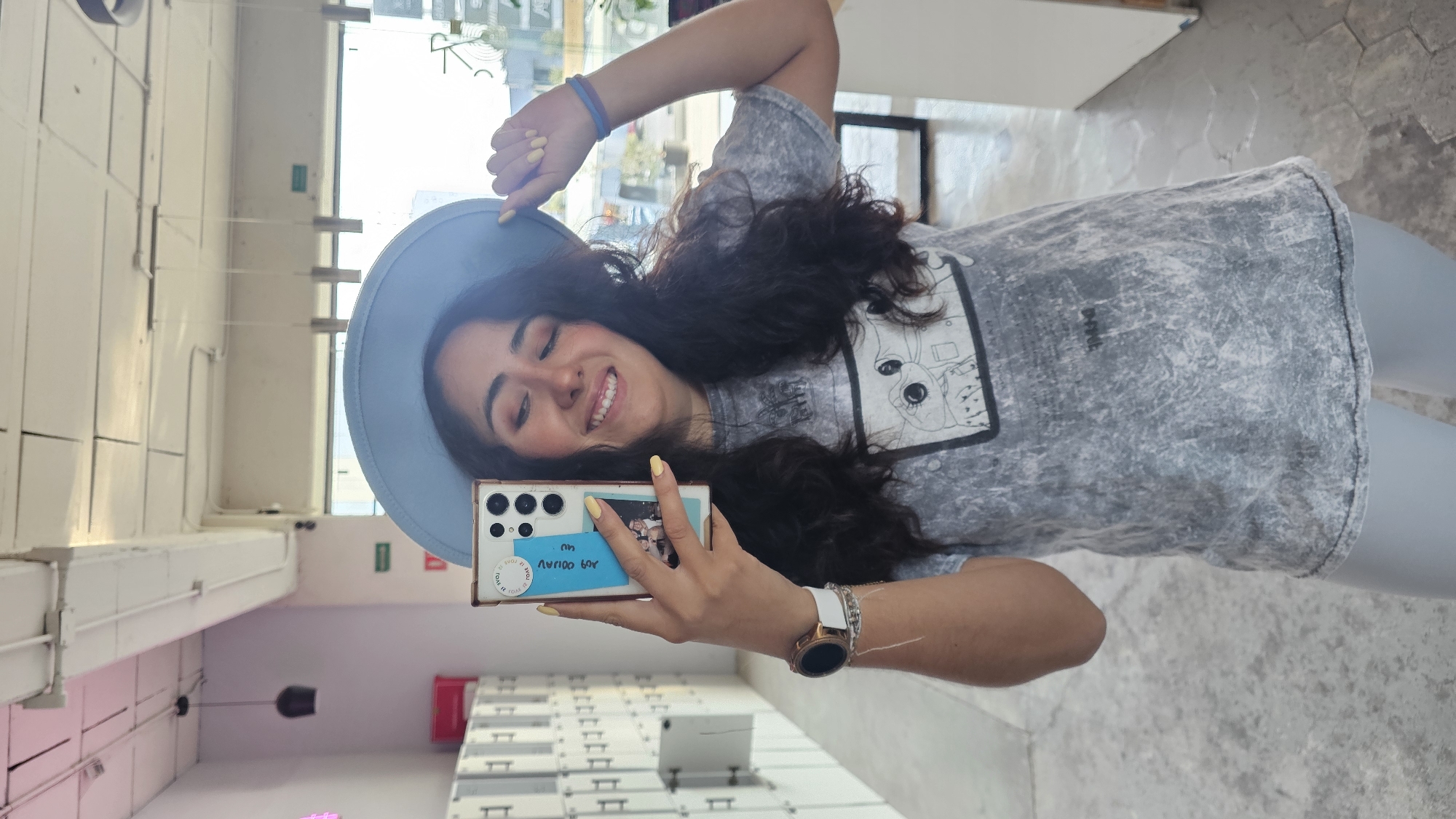 A female is taking a selfie indoors. She is wearing a grey t-shirt, a blue hat and a watch on her left wrist. The location appears to be Puebla, Mexico, based on the locker room-style storage units in the background. The woman is smiling while holding a phone with a picture of 3 people on its case.