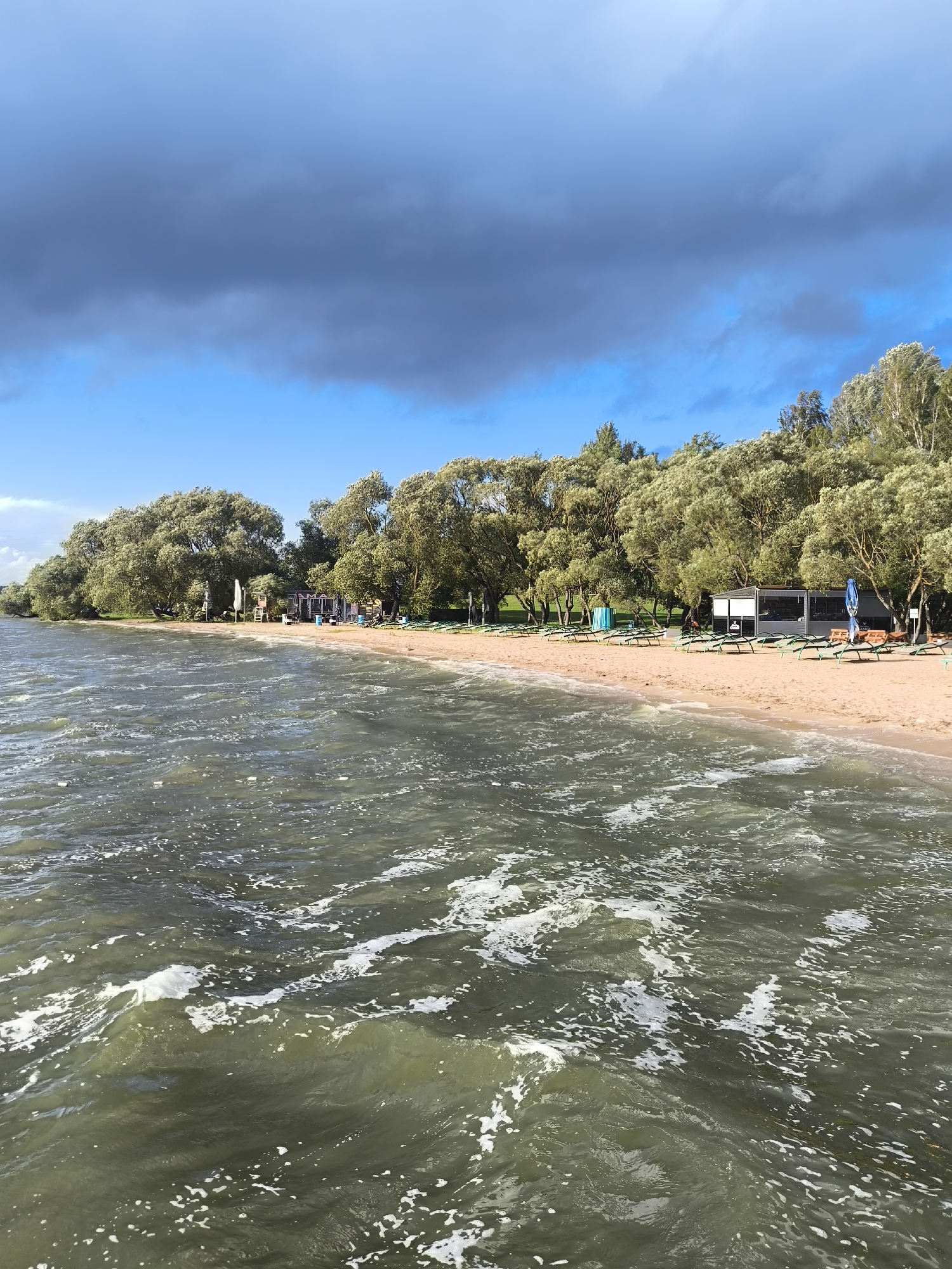 The image depicts a beach scene on a body of water with a sandy shore lined with trees. The foreground shows choppy water with white foam. The beach has several rows of empty lounge chairs and some small structures. The background is dominated by lush green trees, and the sky above is a mix of bright blue and dark, brooding clouds. The location is identified as Ждановіцкі сельскі Савет, Belarus. There are no people visible in the image.