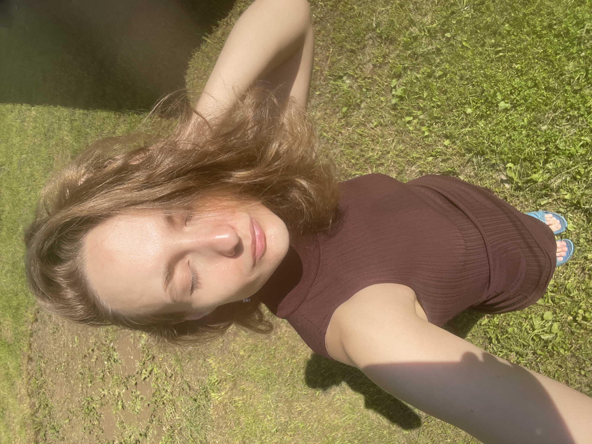 A young woman with light brown hair and closed eyes is captured in a high-angle, selfie-style photograph. She is wearing a dark brown, sleeveless, high-neck ribbed dress and blue flip-flops. Her right arm is bent and rests behind her head, while her left arm is out of frame. The woman is standing on a grassy area with patches of dry earth, suggesting an outdoor environment. The lighting indicates it is a sunny day, casting a shadow on the ground to her left. The background is largely out of focus, with a dark, vertical shadow on the right side of the frame, possibly from a building or structure. The scene appears to be a residential or park area, consistent with a location in Sharkan, Russia.