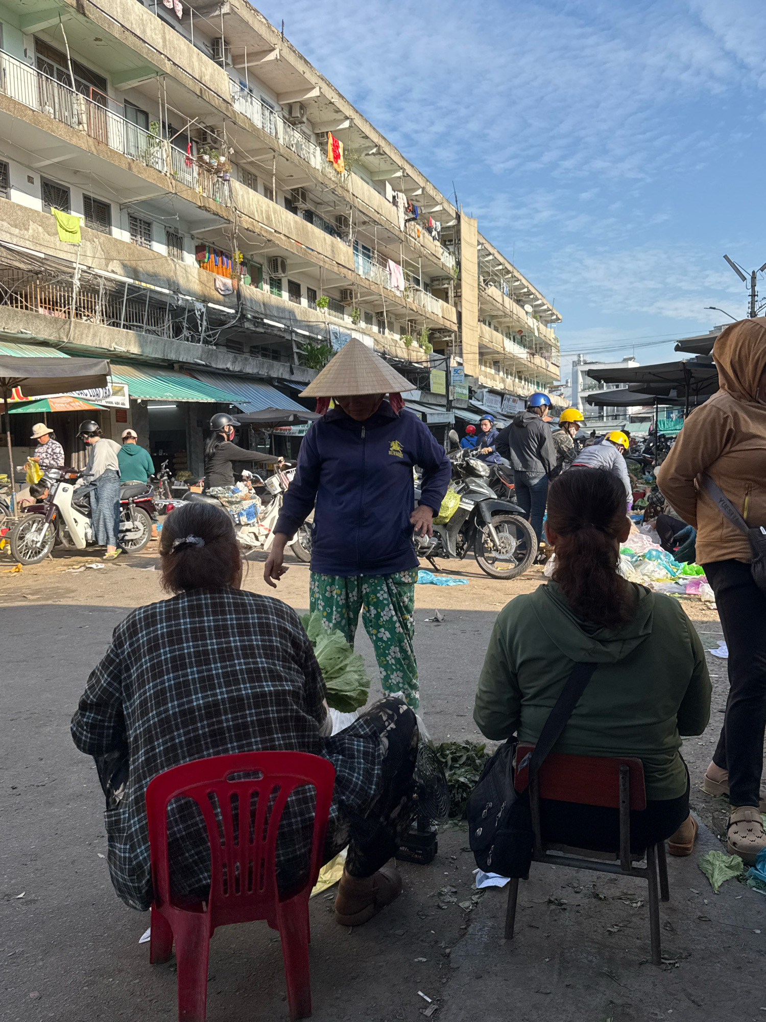 The image depicts an outdoor market scene in Nha Trang, Vietnam, with a multi-story apartment building as a backdrop. The building's exterior is aged, with visible balconies, laundry hanging, and air conditioning units. In the foreground, three individuals are seated on chairs or squatting, facing away from the camera. One person, wearing a plaid shirt, is seated on a red plastic chair. Another, in a green hoodie, is seated on a metal chair with a wooden back. The third individual is partially visible.
Several people on motorbikes are present in the middle ground, suggesting transportation and activity within the market. Some individuals are wearing conical hats and helmets. Various market stalls with awnings are visible, with goods and produce potentially displayed. The ground is paved and appears somewhat cluttered with debris and plastic bags, typical of a bustling open-air market. The sky above is partially cloudy.