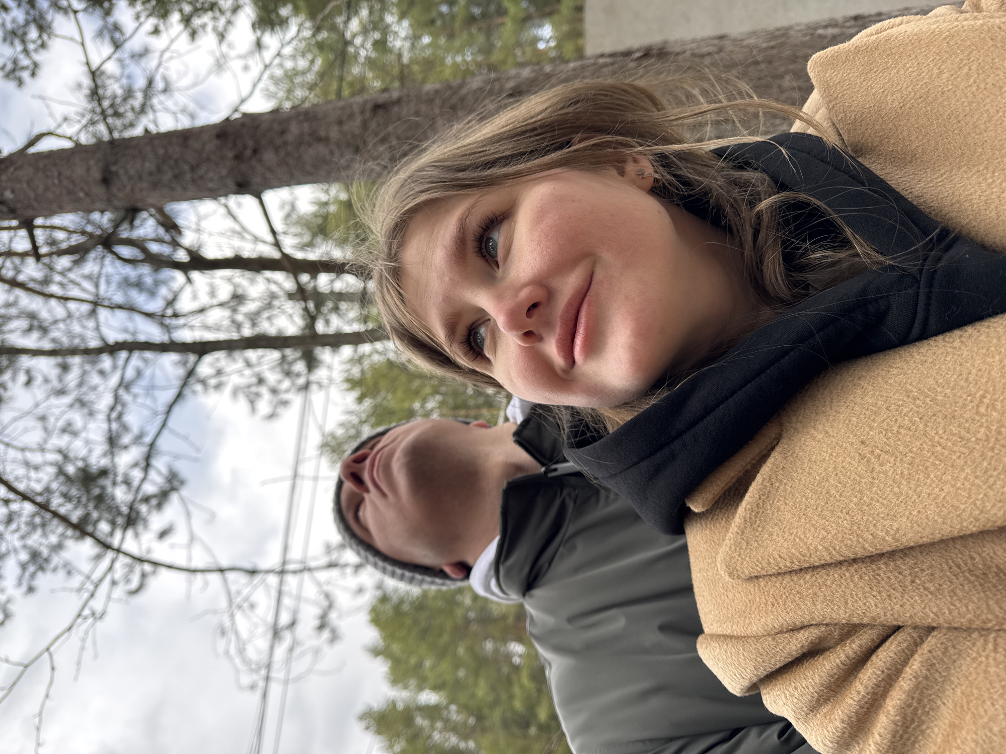 This media file captures two individuals outdoors in Polazna, Russia, seen from a low, upward-looking angle. The primary subject is a woman with light brown hair, fair skin, and blue eyes, positioned in the foreground on the right. She has a subtle, pleasant smile and is looking upwards and slightly to the left. She is wearing a tan-colored coat and a black garment underneath, possibly a scarf or hoodie. A small, star-shaped earring is visible in her left ear.
Partially visible in the background, out of focus and to the left of the woman, is a man wearing a grey beanie, a grey jacket, and a white undershirt. He is also looking upwards.
The scene is set in what appears to be a natural environment, likely a park or wooded area, with numerous trees and branches against an overcast sky. A prominent tree trunk extends across the upper left portion of the image. The sparse foliage on some trees suggests late autumn, winter, or early spring. The lighting indicates it is daytime, possibly a cloudy day given the diffused light and lack of harsh shadows. There is no visible text in the image.