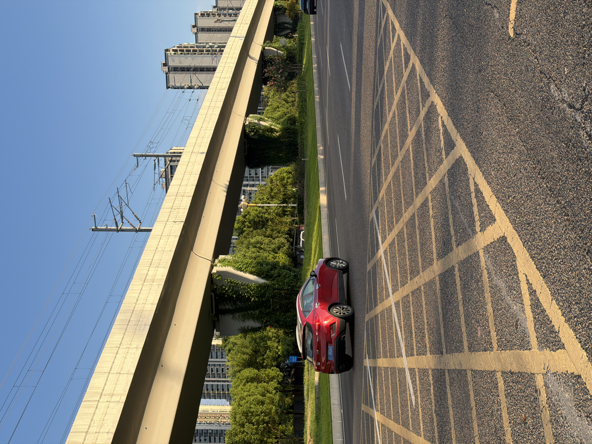 The image shows a red SUV driving on a road under a skybridge in Jiangjin District, Chongqing, China. The skybridge, made of concrete, carries what appears to be a light rail or elevated train line, indicated by the overhead wires and support structures. The road is asphalt with yellow and white lane markings, including a criss-cross yellow box marking in the foreground. Lush green trees line the side of the road, and several modern high-rise buildings are visible in the background. The sun is low in the sky, casting long shadows and illuminating the scene with warm light, suggesting it is late afternoon or early evening. The sky is clear and blue.