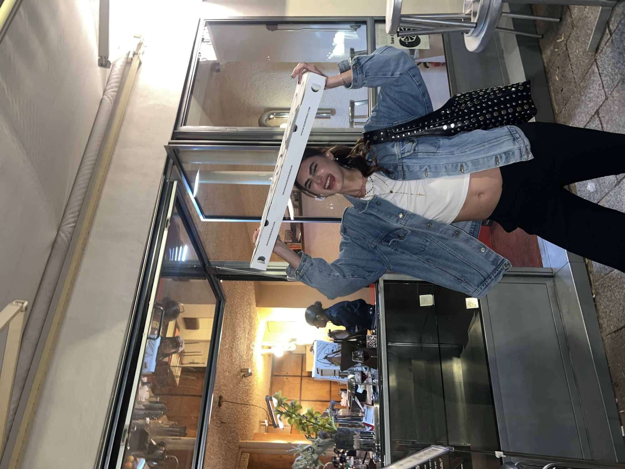A woman is standing outside a restaurant. She is holding an open pizza box above her head. She is wearing a denim jacket, white top, and black pants. The restaurant has large windows that reflect the interior. There are tables and chairs inside the restaurant, and a person can be seen working behind the counter. The image was taken in Paris, France.