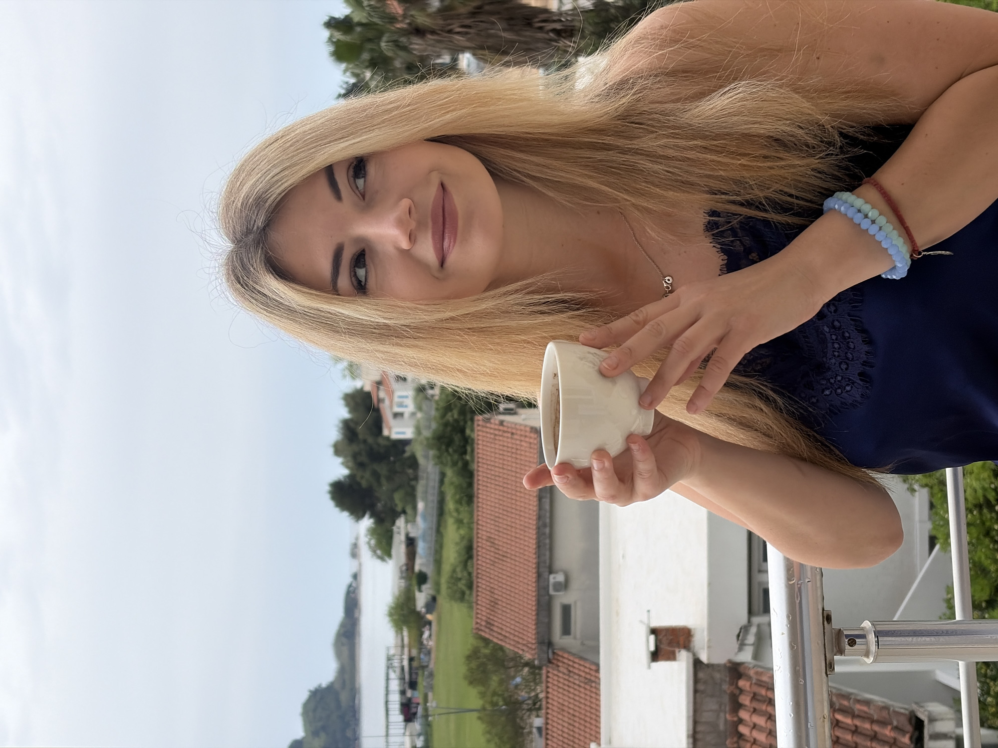 A young woman with long, straight blonde hair, fair skin, and a light smile is depicted in a medium close-up, looking directly at the camera. She wears a dark blue top, possibly with delicate lace trim on the shoulder, and a silver necklace with a small charm. On her left wrist, she sports two beaded bracelets—one light blue and one light green—alongside a thin red string bracelet. She holds a white ceramic mug in both hands, positioned near her chest.
The setting is outdoors, likely a balcony or terrace, indicated by a metal railing visible on the right. The background features a charming coastal town, consistent with Budva, Montenegro. Several buildings with distinct reddish-orange tiled roofs and white walls are visible, interspersed with lush green trees and foliage. Beyond the town, a calm body of water, possibly a bay or the sea, extends towards a distant landmass under a bright, clear sky. The scene suggests a relaxed moment during the daytime, enjoying a beverage in fair weather within a picturesque Mediterranean environment. No text is visible in the image.