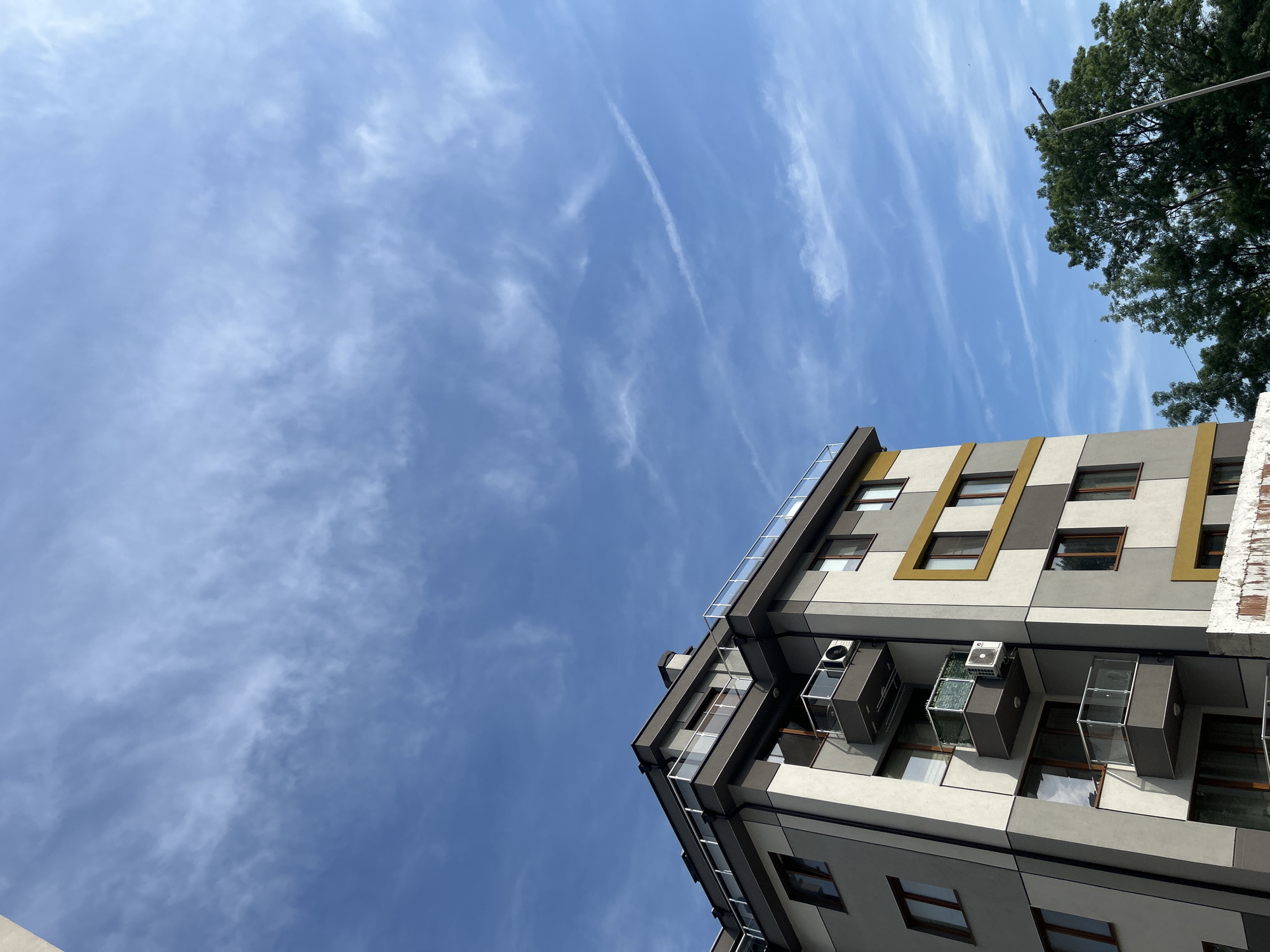 The image displays a modern, multi-story residential building under a clear blue sky sparsely populated with wispy cirrus clouds and a single contrail in the upper right. The building's exterior is characterized by a combination of light gray and off-white facade panels. Several windows are highlighted by rectangular yellow-gold frames. Multiple balconies project from the structure, each featuring clear glass and metal railings. Visible on these balconies are two outdoor air conditioning units; one balcony also incorporates a green mesh screen. The building's top edge includes a similar glass and metal railing, suggesting a rooftop or penthouse terrace. In the lower right quadrant, dense green deciduous tree foliage is visible, partially obscuring a utility pole or street light. The bottom edge of the frame reveals a section of a rough, unfinished concrete structure or wall. The scene is illuminated by direct natural daylight. This architecture is characteristic of contemporary urban development in residential areas within the city of Varna, Bulgaria.