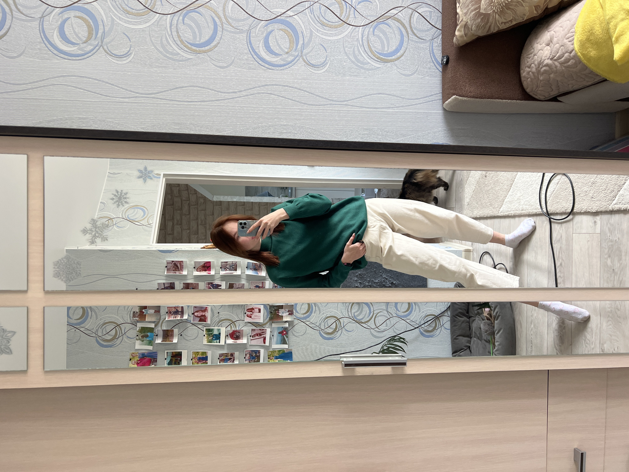 The image displays a full-length mirror selfie of a woman in an indoor setting, likely a home in Chaikovsky, Russia. The woman has reddish-brown hair and is dressed in a dark green long-sleeved top and light beige trousers, paired with white socks. She holds a silver smartphone in front of her face, partially obscuring it, as she captures her reflection.
The mirror, possibly integrated into a wardrobe, is composed of rectangular panels. In the reflection, patterned wallpaper featuring blue and grey swirls is visible on the wall behind the woman. Below her, a light-colored floor, resembling wood laminate, is seen, along with a curled black electrical cord. A tabby cat is also visible on the floor to the right of her legs in the reflection.
Several small, printed photos or polaroids are arranged in multiple rows on the wall or mirror frame behind the woman, showcasing various people and scenes, lending a personalized feel to the space. A small potted plant is also reflected in the lower mirror segment.
Outside the mirror's reflection, the same patterned wallpaper is visible on the upper wall. To the right, a section of a sofa or bed with several cushions in brown, beige, and yellow tones is present. The lower part of the image shows the light wooden structure of a wardrobe or cabinet to which the mirror is attached. The scene is casual and domestic, likely captured during the daytime. No discernible text is visible.
