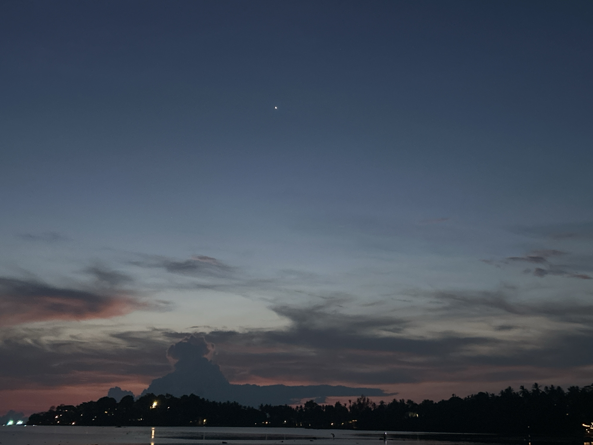 The image shows a twilight scene in Baan Sri Thanu, Thailand. The sky is a gradient of colors, ranging from dark blue at the top to a dusky orange and pink near the horizon. Clouds are present, some thin and wispy, others thicker and more defined. A single bright point of light, possibly a star or planet, is visible in the upper part of the sky. Below the sky, a line of dark trees and vegetation sits on what appears to be a shoreline. Small points of light dot the landscape, likely from buildings or other structures. A body of water, possibly the sea or a lagoon, is visible in the foreground.