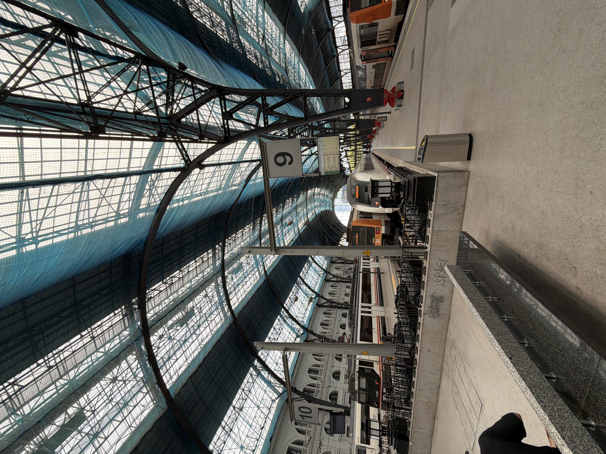The image shows a view of the roof and platforms of a train station in Barcelona, Spain. The roof is a large, arched structure made of metal and translucent material, with blue netting covering sections of it. The station platforms are wide and empty, with trains parked at some of them. The architecture of the station building in the background is classical, with arched windows and decorative elements. In the foreground, there is a granite bench and a trash can. There are no people visible in the image.