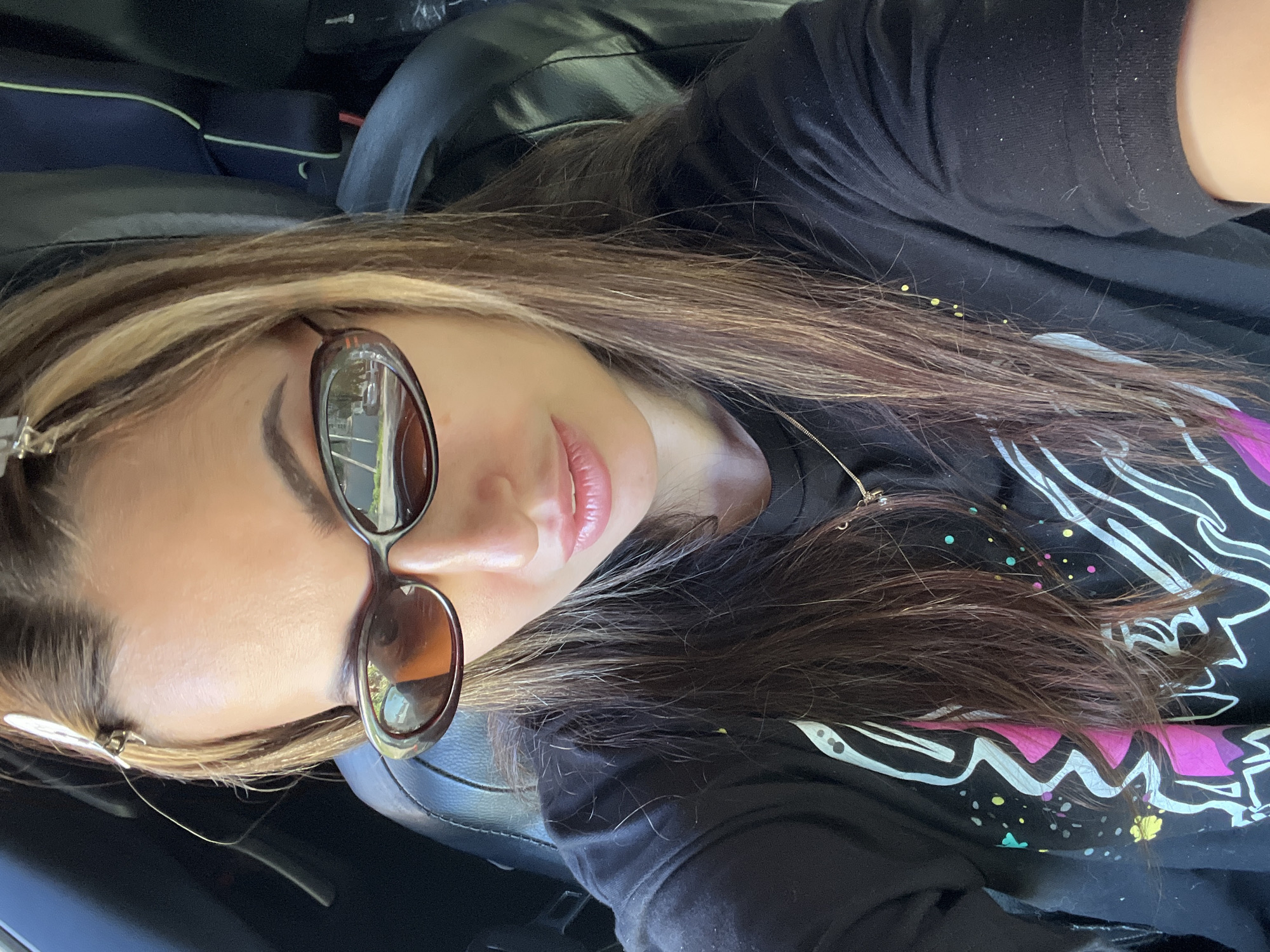 A medium-close-up, low-angle shot shows a woman wearing sunglasses and a black graphic t-shirt in a car. The woman is looking slightly down at the camera with a neutral expression. She has long brown hair with blonde highlights, held back by a clip. She is wearing sunglasses with brown frames and tinted lenses. The t-shirt features a white and pink graphic design on the front. The car's interior is visible in the background. The location is Unknown, Unknown.